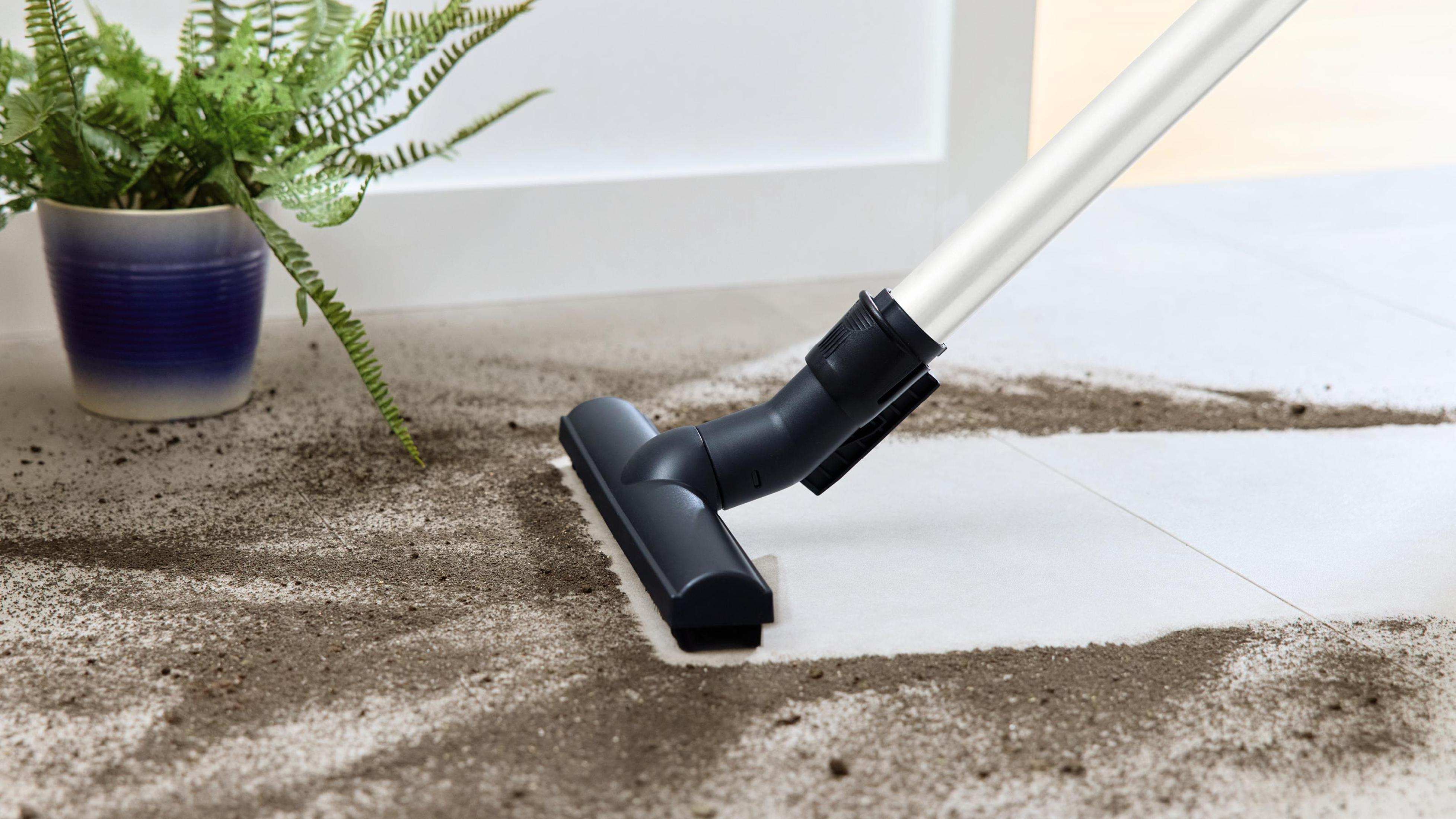 Głowica odkurzacza na białej podłodze z brudem i rośliną doniczkową w tle, czyszcząca podłogę.