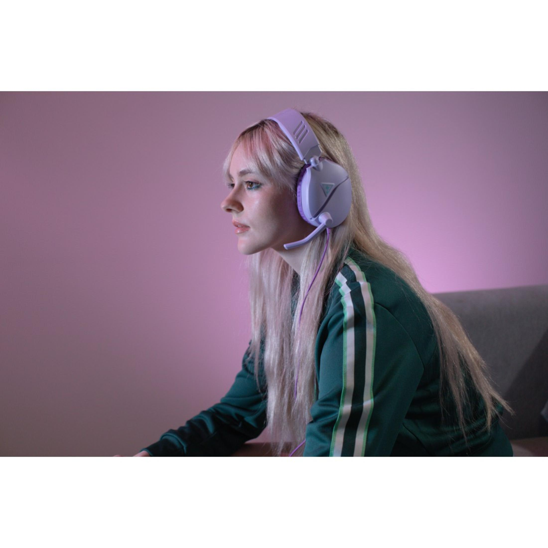 Kobieta z blond włosami i zestawem słuchawkowym gra. Tło jest różowe, z zielono-białą kurtką w paski.