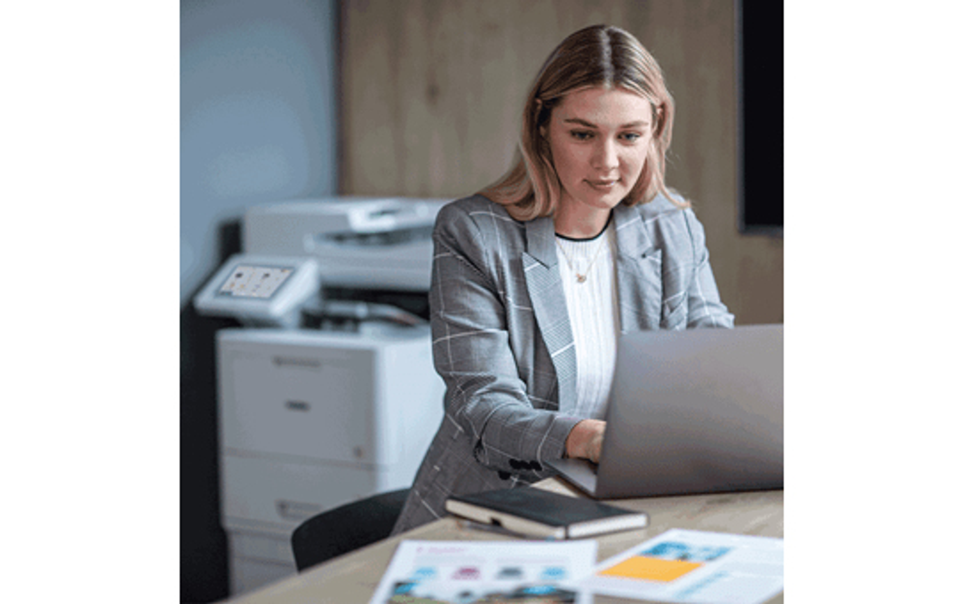Kobieta w szarej marynarce pracuje na laptopie. Drukarka widoczna w tle.