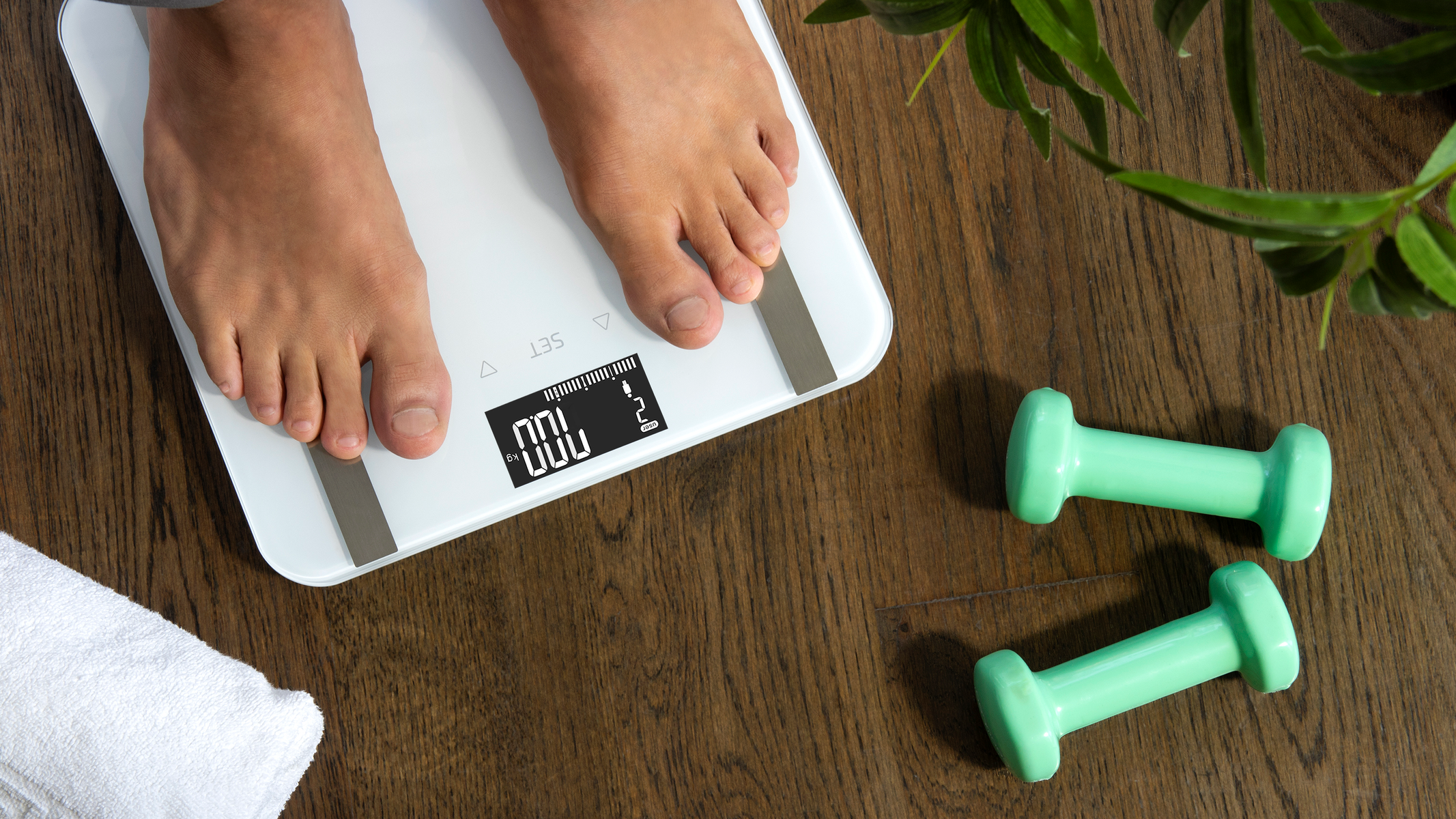 Stopy na białej wadze, z ciężarkami i rośliną na drewnianej podłodze.