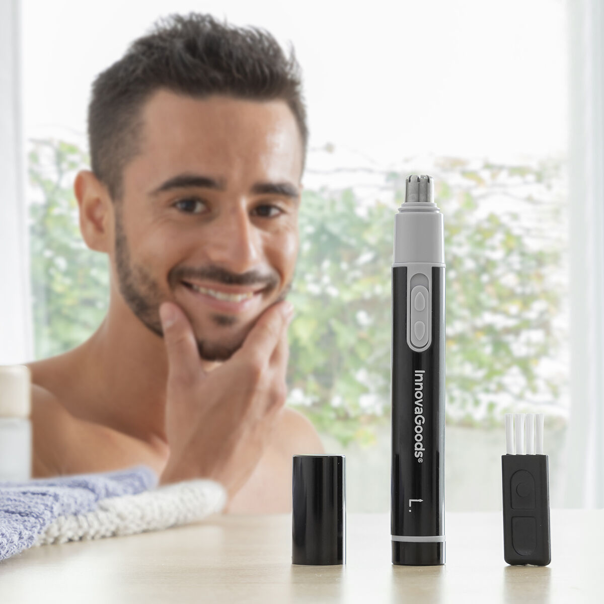 Hombre sonriendo, sosteniendo su cara. Cortapelos nasal y accesorios sobre la mesa. Entorno de baño brillante.
