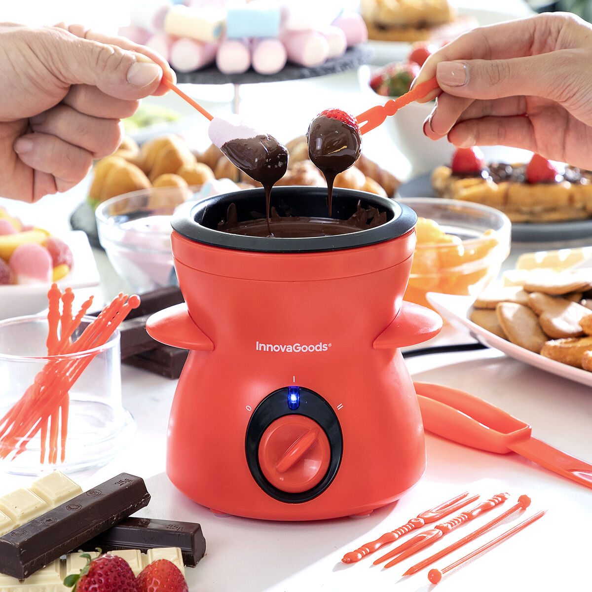 Ein roter InnovaGoods Fondue-Topf mit Schokolade und Erdbeeren, jemand taucht Marshmallows ein.