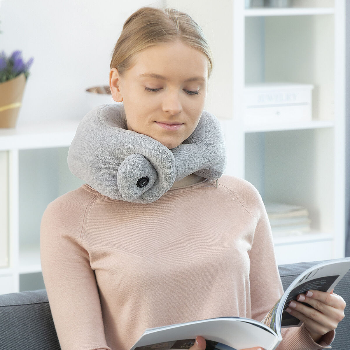 Una mujer usando una almohada para el cuello gris y leyendo una revista en un sofá.