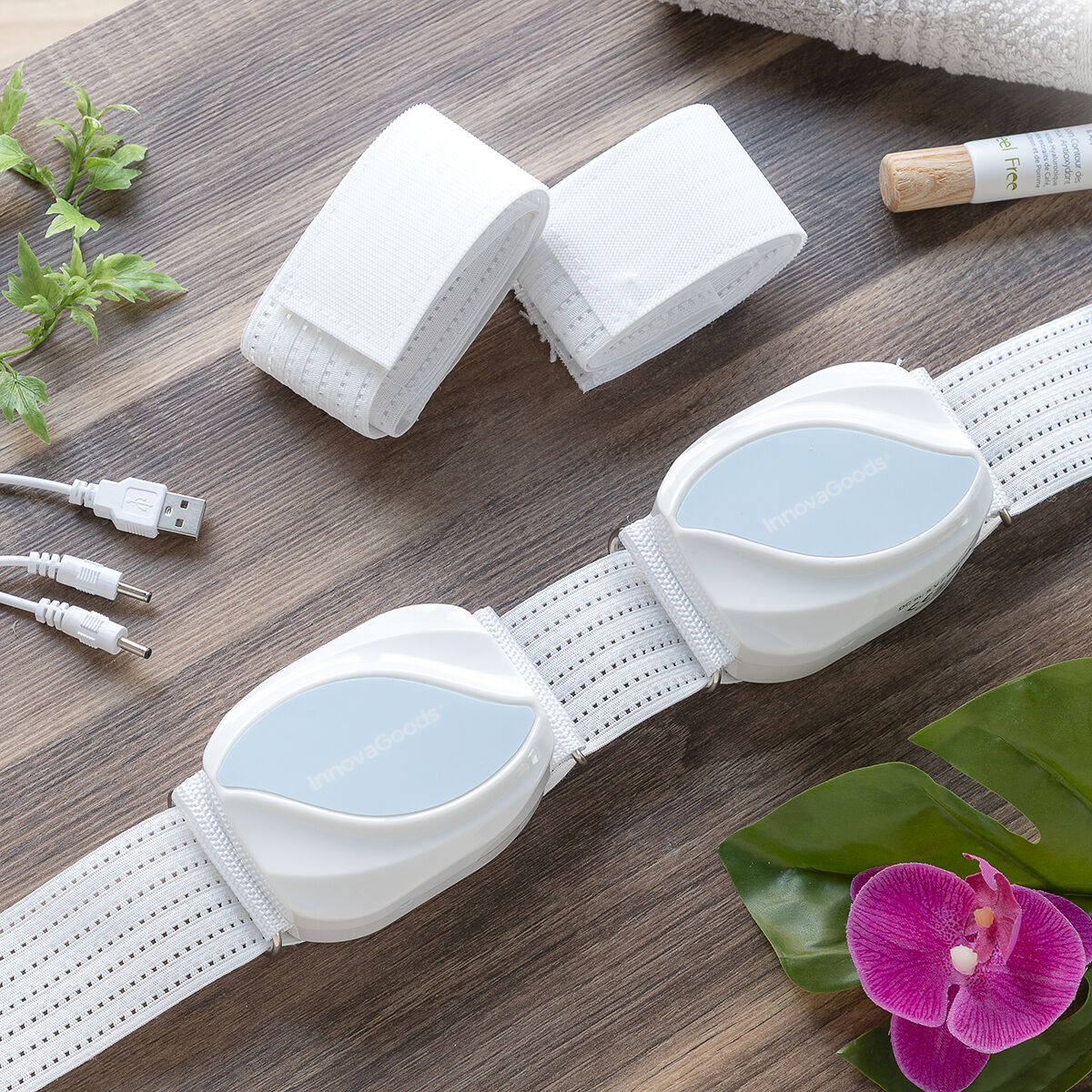 Un cinturón de masaje blanco con dos módulos InnovaGoods azules, sobre mesa de madera, con hojas y flor.