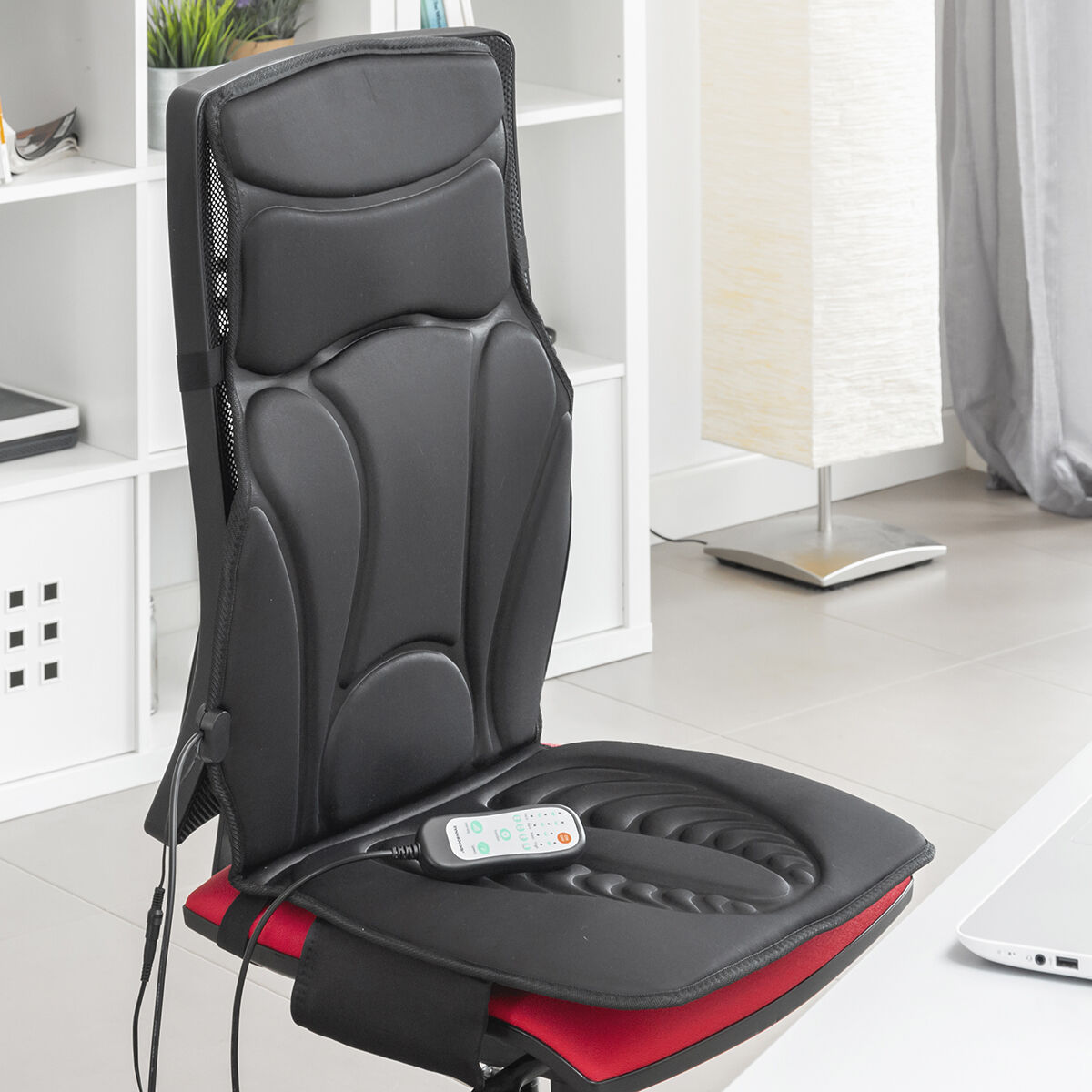 Un cojín de masaje negro para silla en una silla roja, con un mando a distancia. Hay una lámpara y una estantería blanca al fondo.