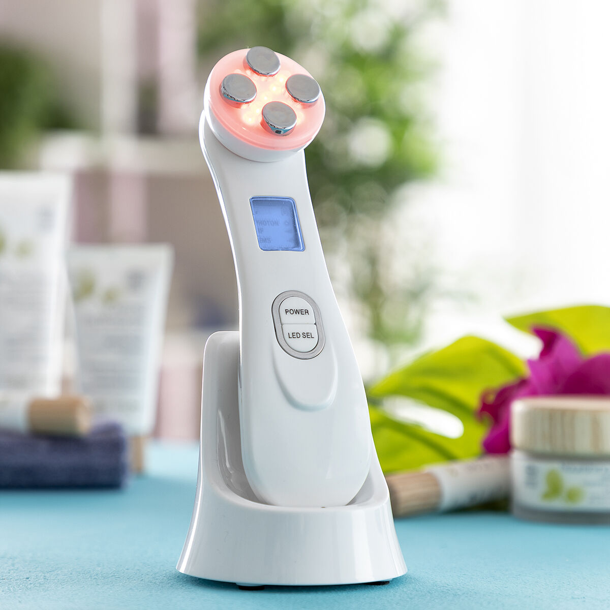 Dispositivo facial blanco con cuatro luces. Base de carga abajo. Artículos cosméticos en la superficie azul.