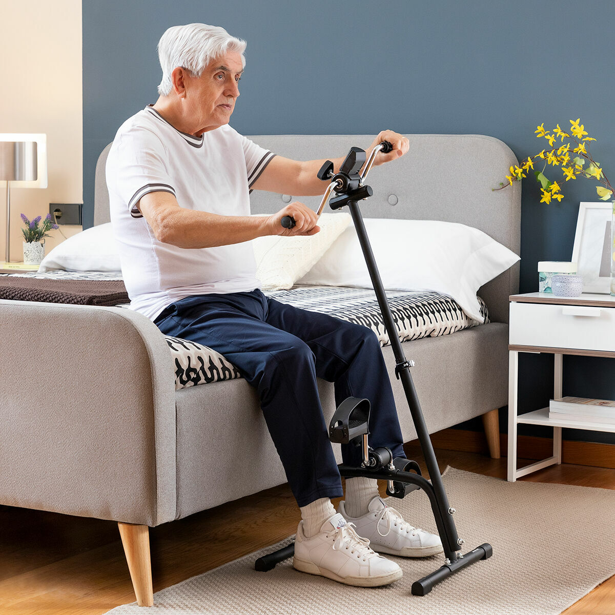 Ein älterer Mann benutzt eine Bein-Trainingsmaschine, er sitzt auf einem Bett. Im Hintergrund ein Raum.