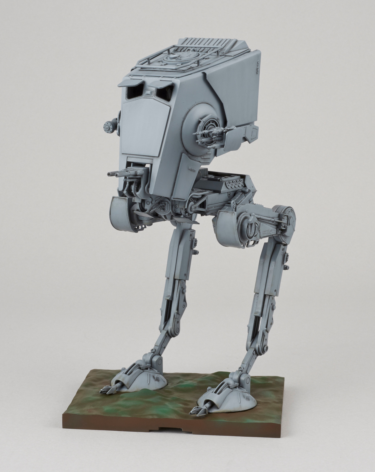Un modèle gris d'AT-ST de Star Wars se dresse sur une base verte et marron. Le fond est gris.