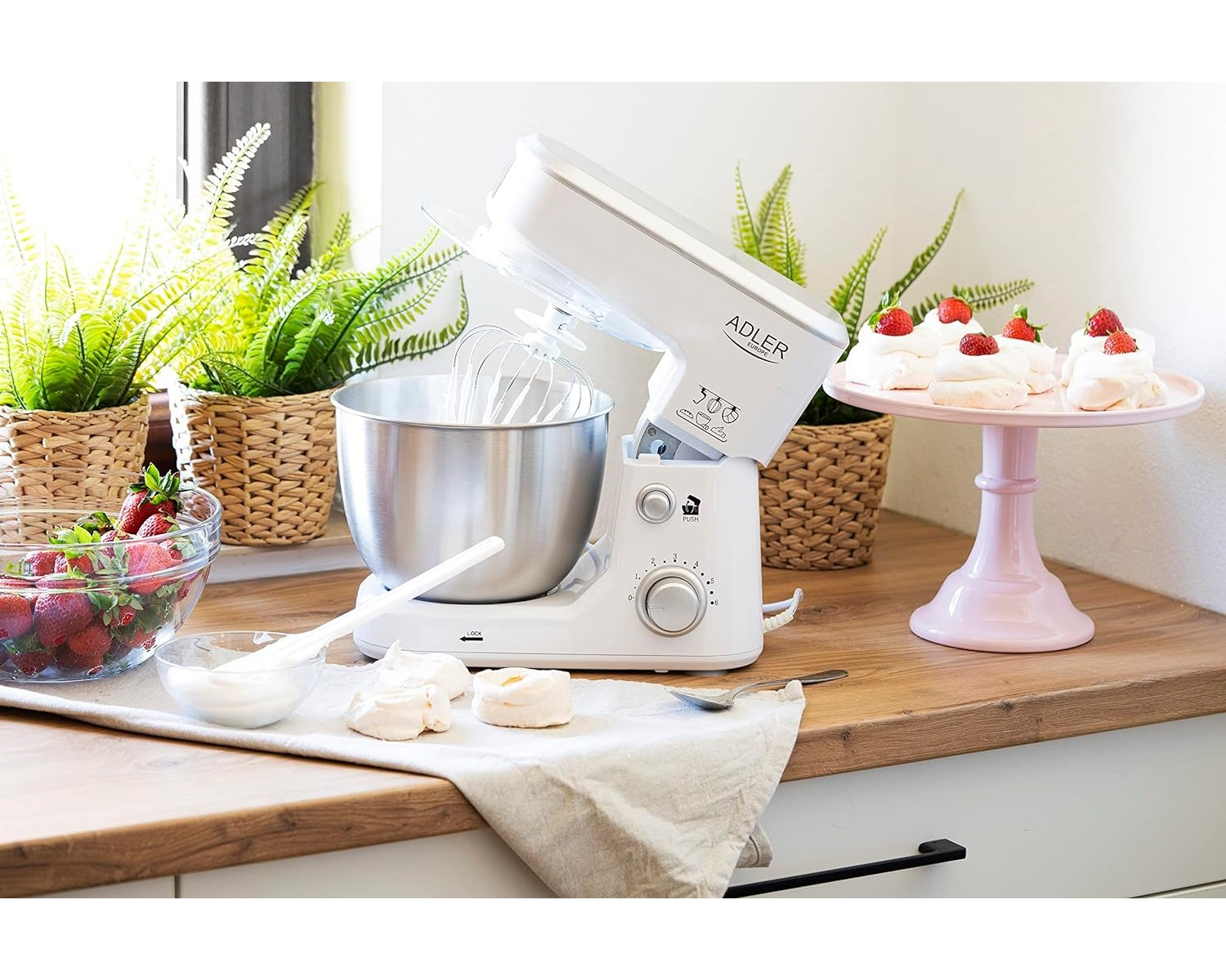 Weißer Küchenmixer mit Schüssel, Schneebesen und Baiser. Erdbeeren und Pflanzen im Hintergrund.
