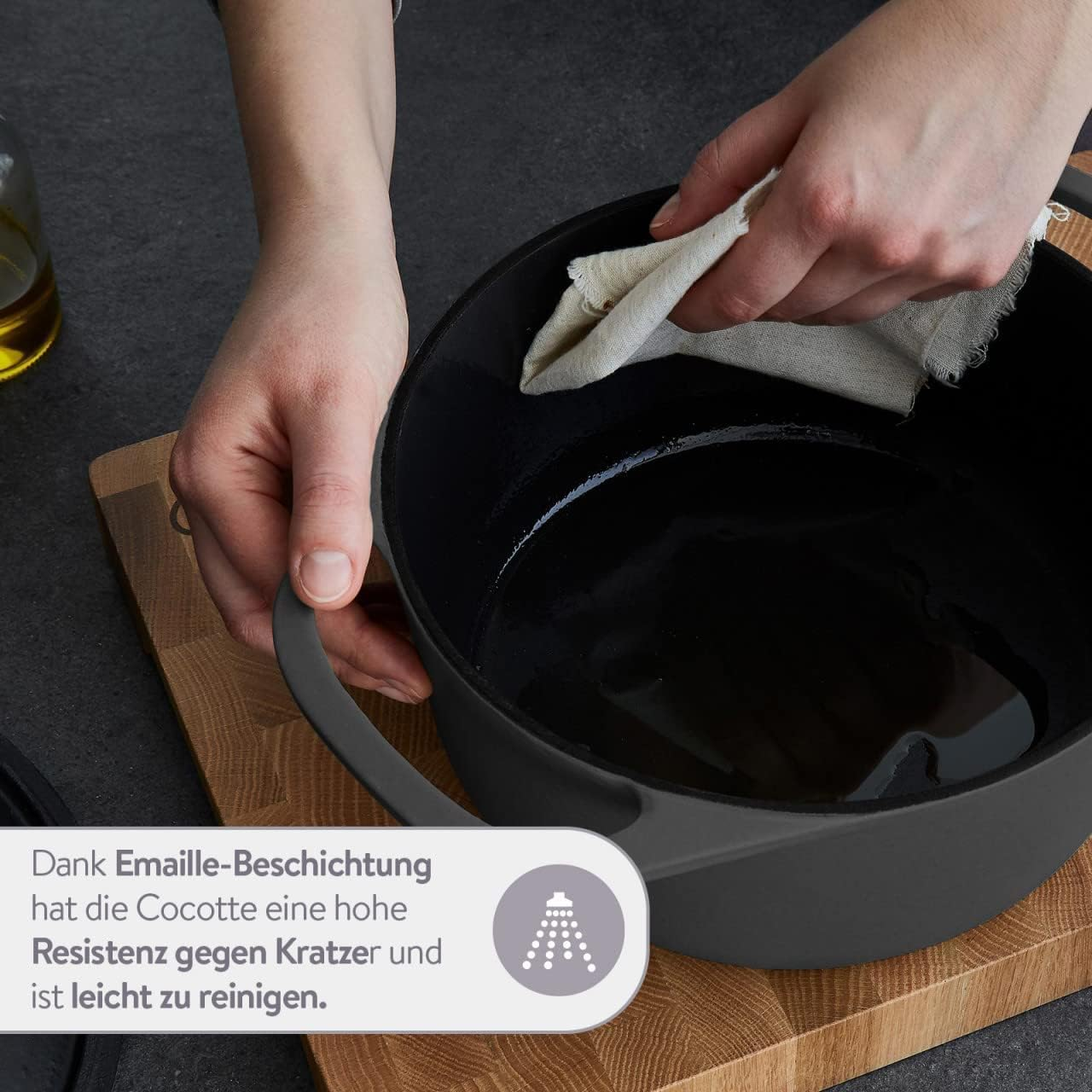 Hände reinigen einen schwarzen Topf mit einem Tuch. Der Topf steht auf einem Holzschneidebrett auf dunkler Oberfläche.