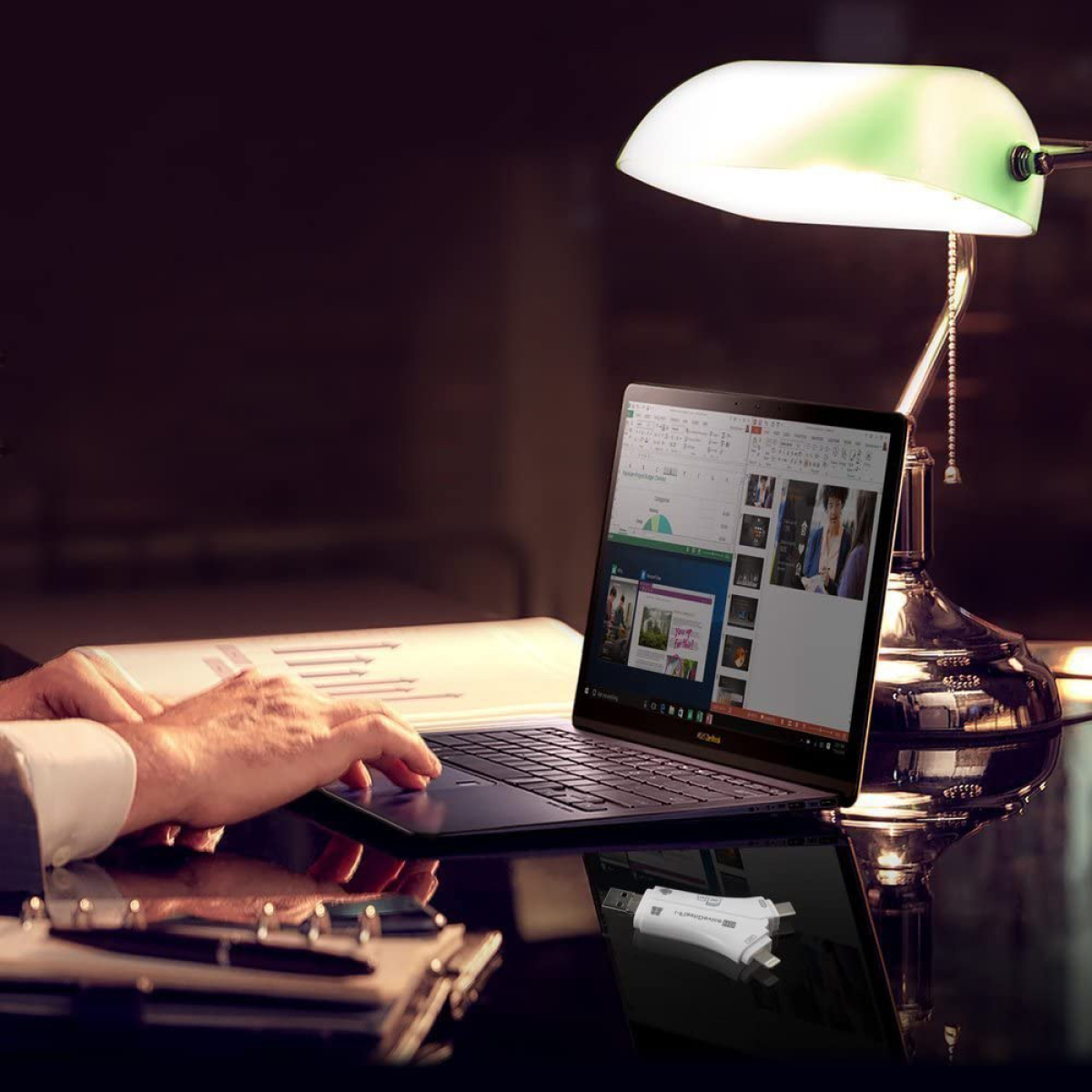 Ein Laptop auf einem Schreibtisch mit Lampe und Händen. Ein USB-Stick liegt da.