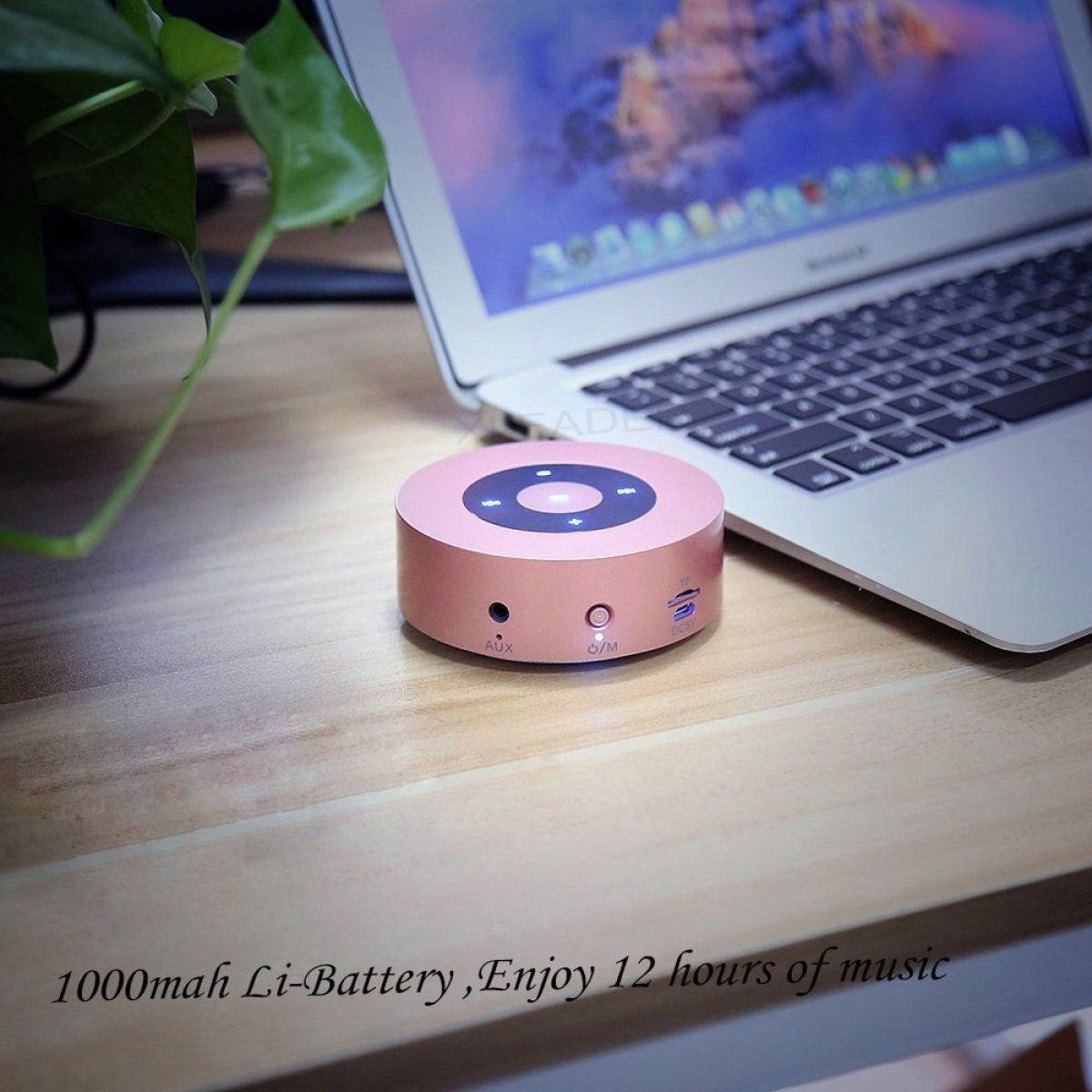 Ein kleiner rosa Lautsprecher auf einem Holztisch mit einem Laptop im Hintergrund. Text: '1000 mAh Li-Akku, 12 Stunden genießen'.