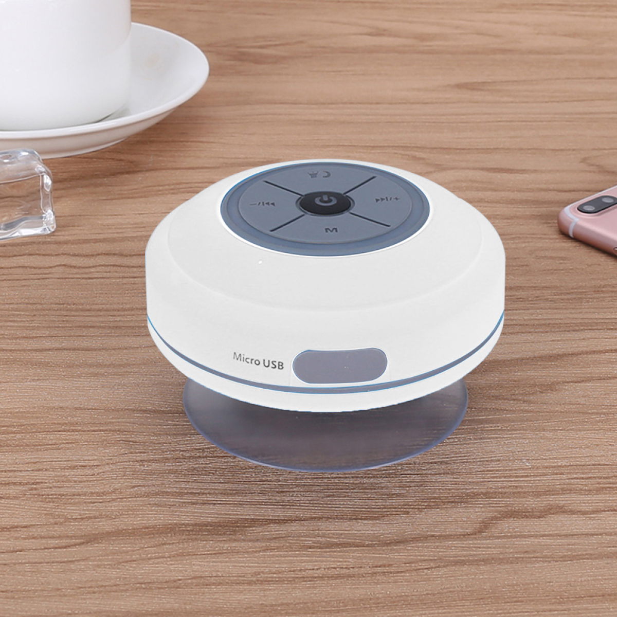 Weißer wasserdichter Lautsprecher mit Saugnapf auf Holzoberfläche, neben einer Tasse und Telefon.