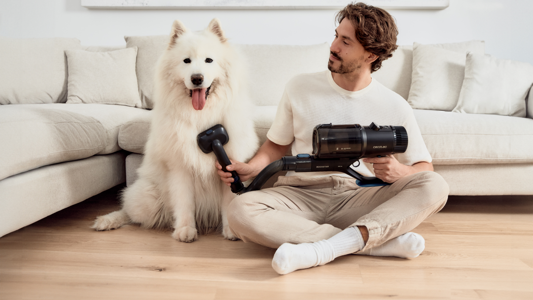 Uomo che aspira un cane bianco seduto su un pavimento di legno chiaro, vicino a un divano color crema.
