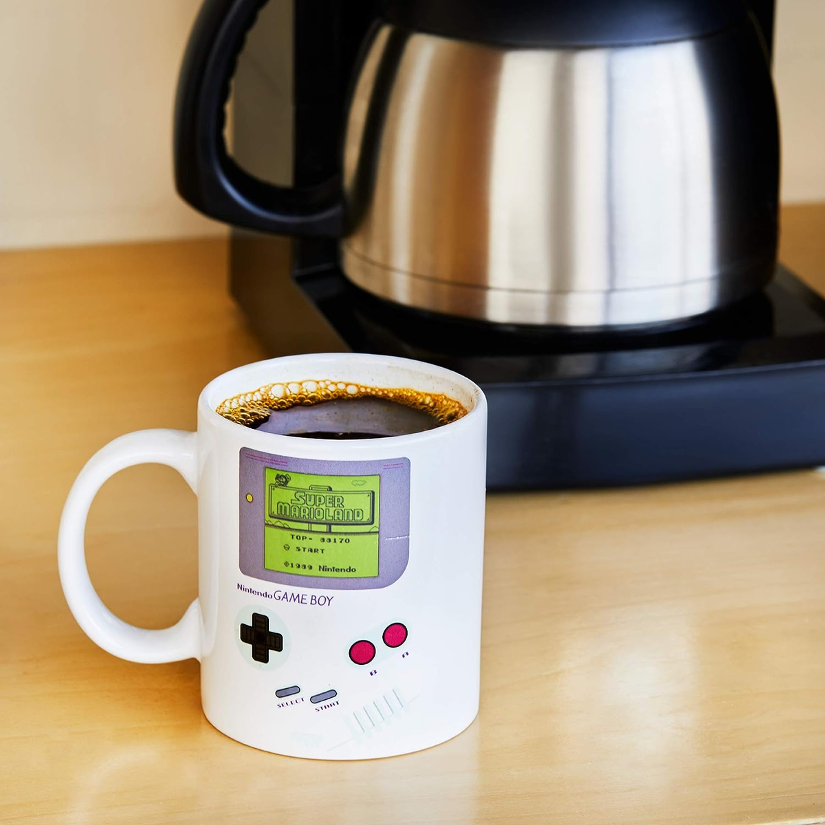 Taza blanca con diseño de Super Mario Gameboy. Café en la taza, cafetera negra detrás.