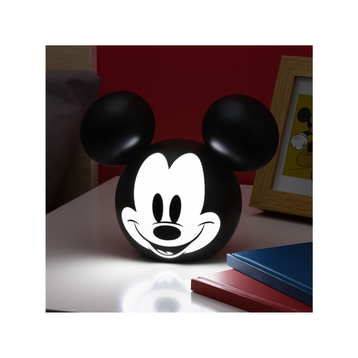 Lámpara de Mickey Mouse. Cabeza negra con cara y orejas blancas, iluminada en mesa blanca. Fondo de pared roja.