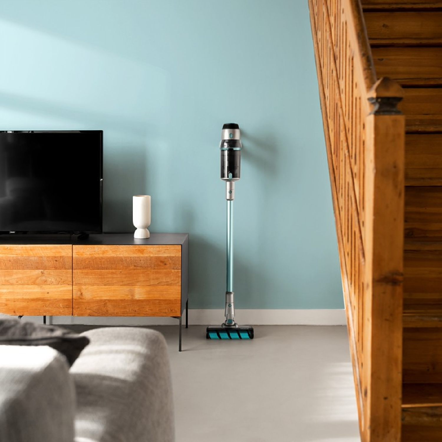 Aspirapolvere senza fili appoggiato a una parete blu. Mobile in legno e scale. Vaso bianco.