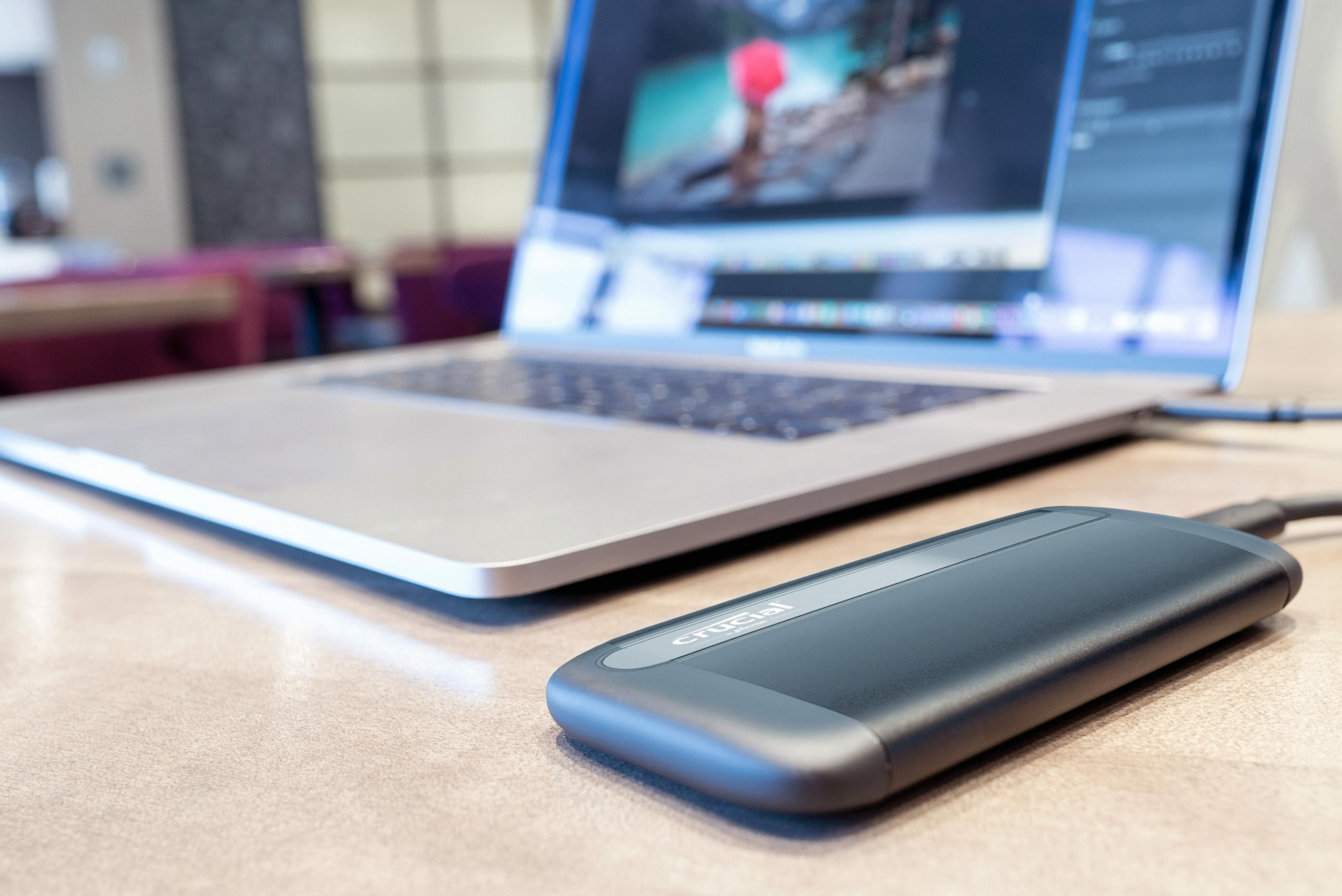 Czarny zewnętrzny dysk twardy, oznaczony jako 'Crucial', jest podłączony do laptopa na biurku.