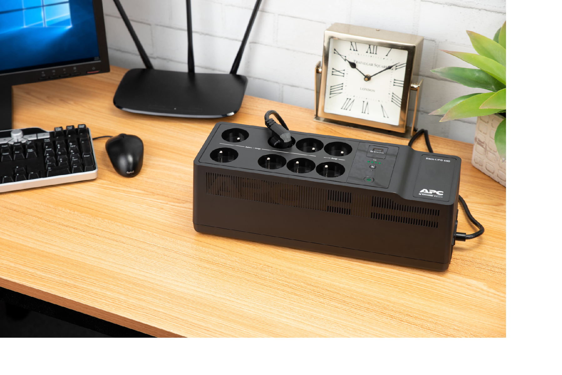 UPS nero con prese, su scrivania in legno. Router, orologio e pianta visibili sullo sfondo.