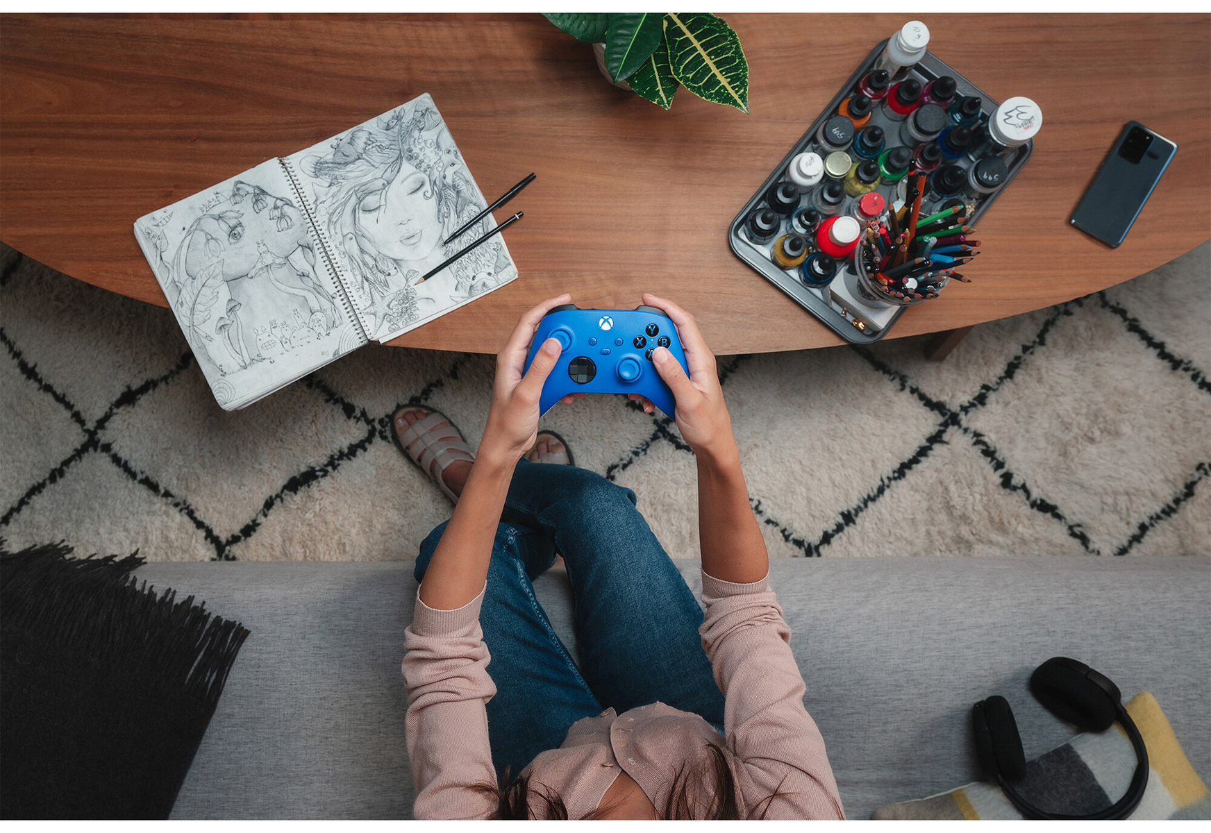 Person hält einen blauen Xbox-Controller auf einem Tisch mit Kunstbedarf und Telefon.