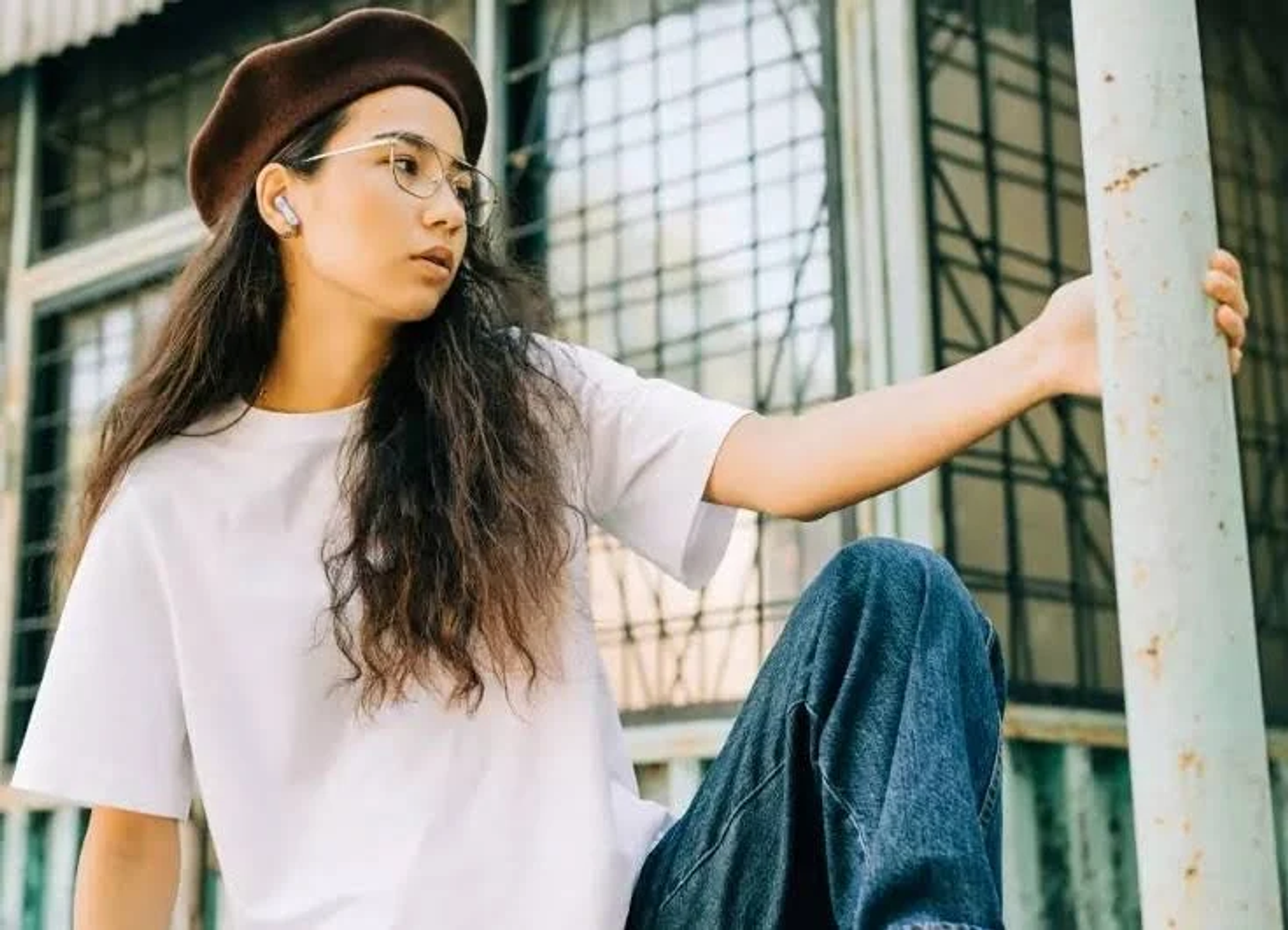 Donna con camicia bianca, jeans e basco, con auricolari, appoggiata a un palo di metallo.
