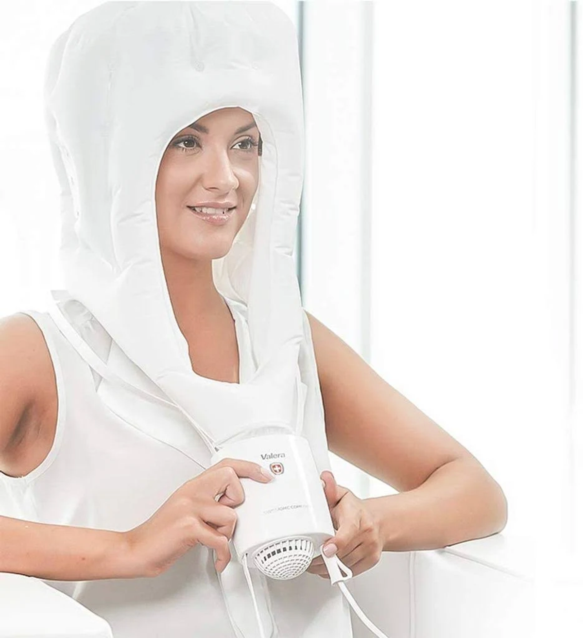 Kobieta w białym kapturze suszarki do włosów, uśmiechnięta, trzymająca suszarkę.