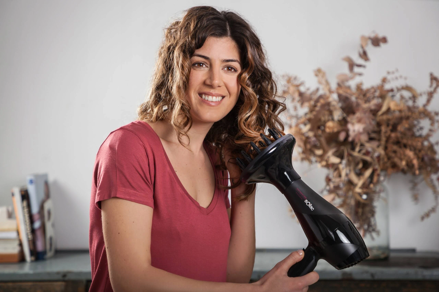 Kobieta suszy włosy czarną suszarką. Uśmiecha się i patrzy w kamerę.