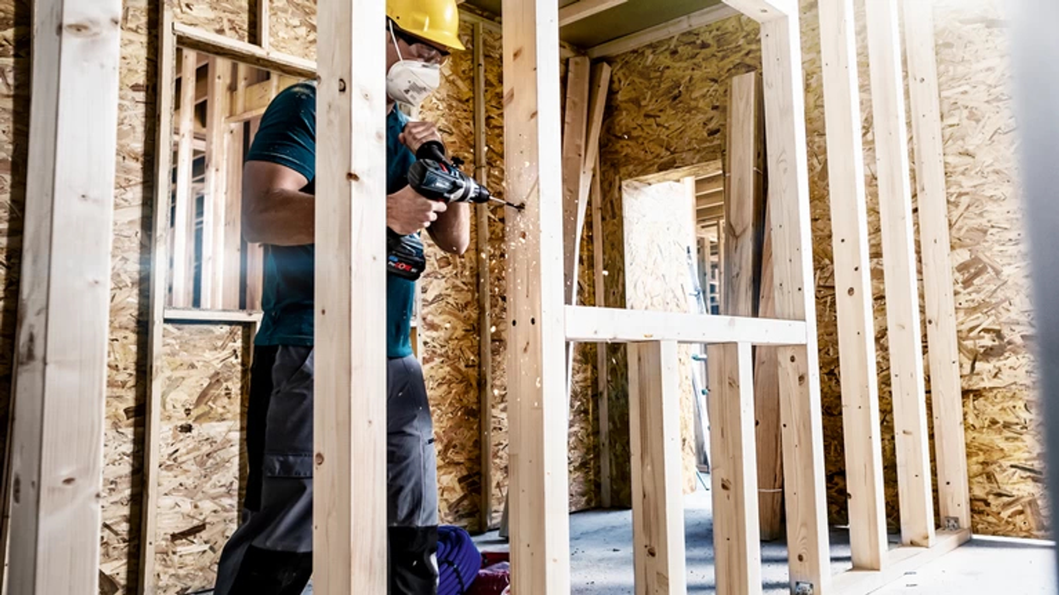 Robotnik budowlany wiercący w drewnianych słupkach, w kasku i masce.