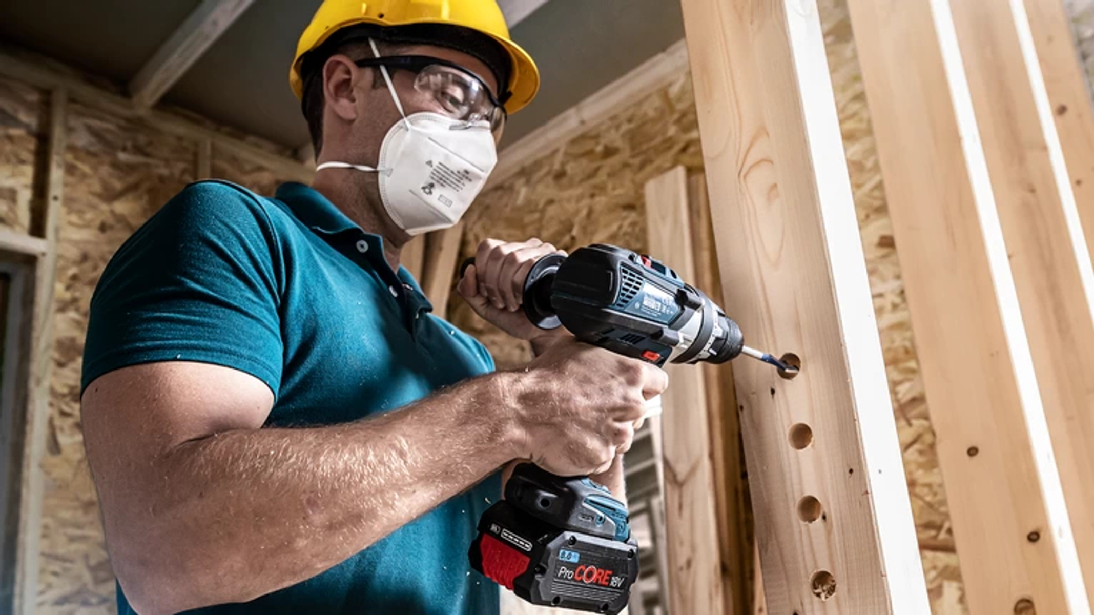 Mężczyzna w masce i kasku wierci w drewnianych słupkach. Wiertło i drewno są w centrum uwagi.