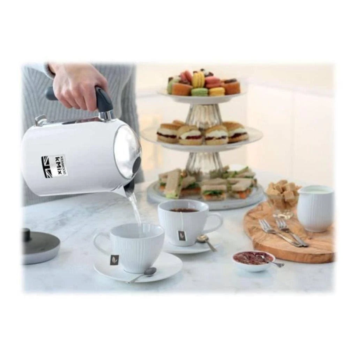Eine Person gießt heißes Wasser aus einem weißen Wasserkocher in eine Teetasse. Tee und Snacks stehen auf einem Tisch.