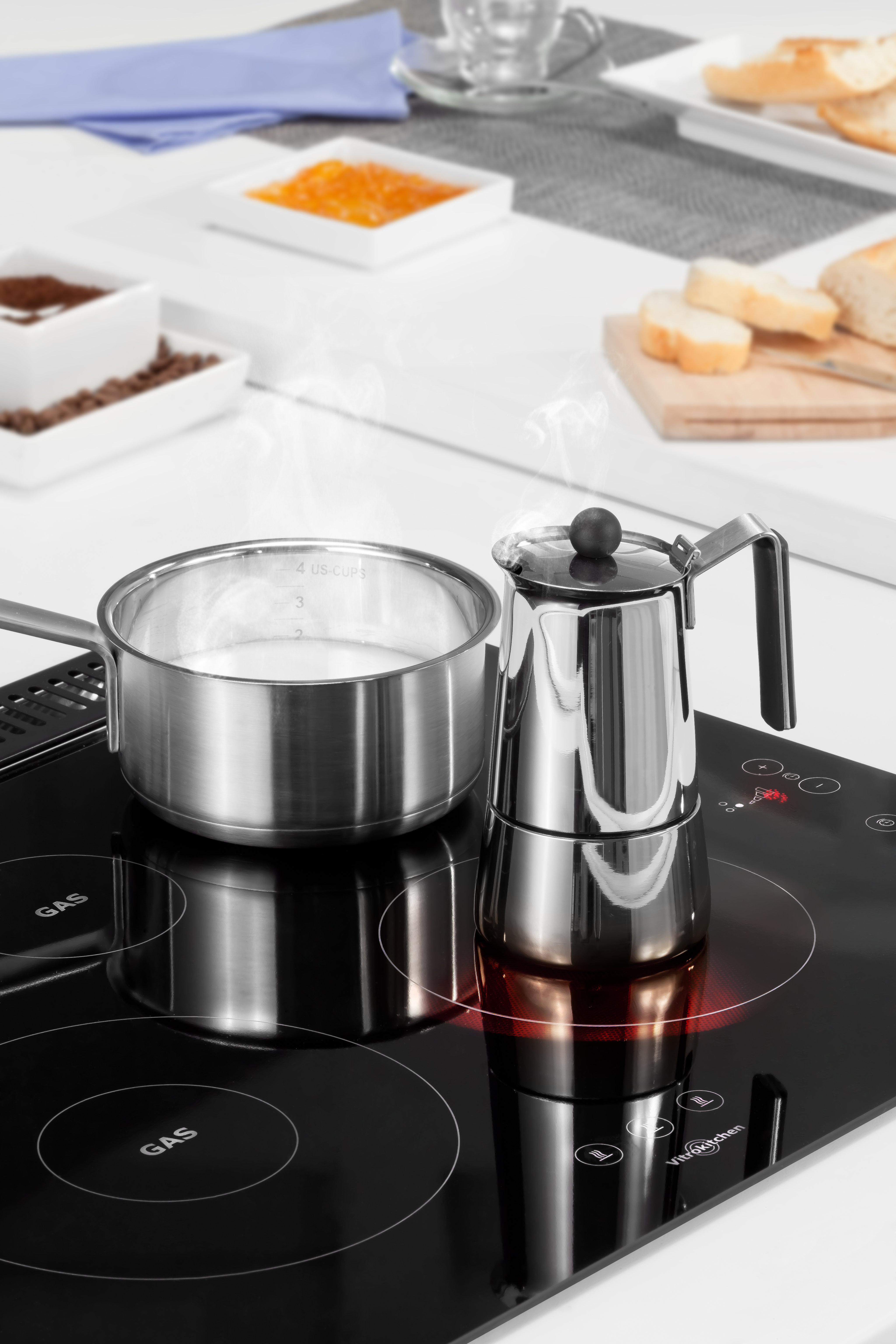 Una vitrocerámica de acero inoxidable con una olla de leche y una cafetera. Pan y mermelada al lado.