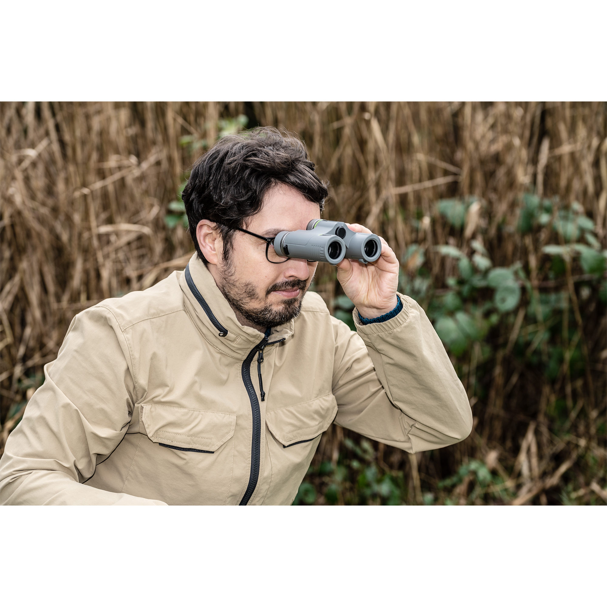Hombre con gafas usando binoculares en un campo. Lleva una chaqueta beige y mira a través de ellos.