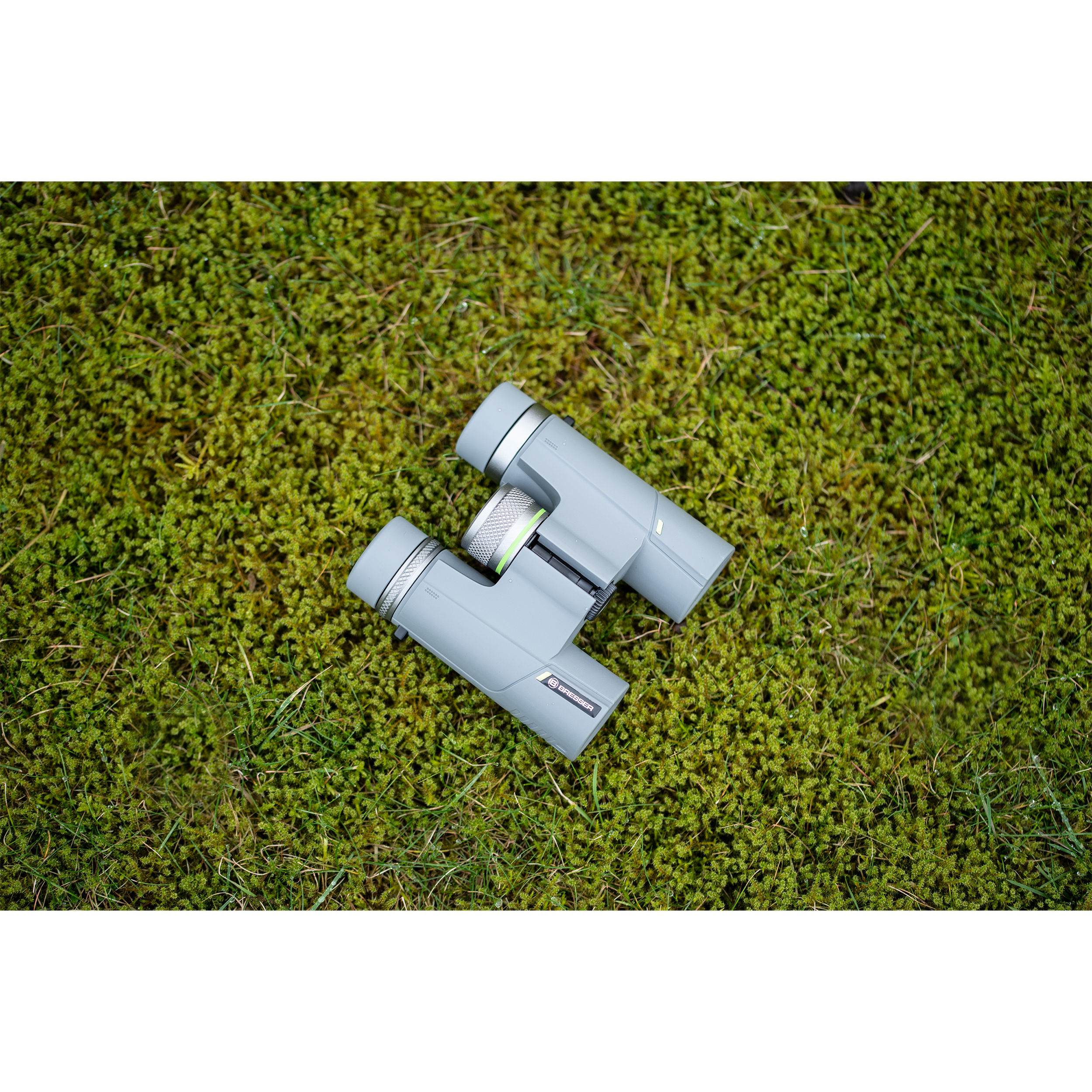 Prismáticos grises sobre musgo verde. Los prismáticos tienen una rueda plateada en el medio.