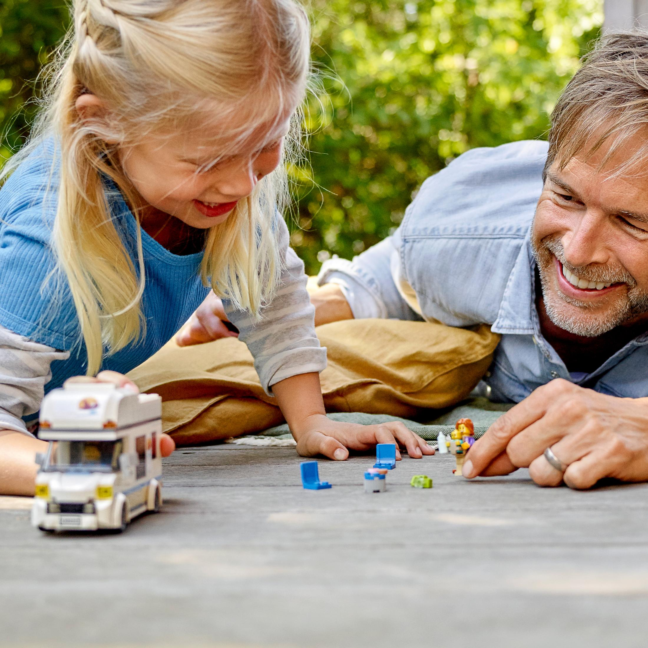Een vader en dochter spelen met een speelgoed camper en figuren op een houten oppervlak.