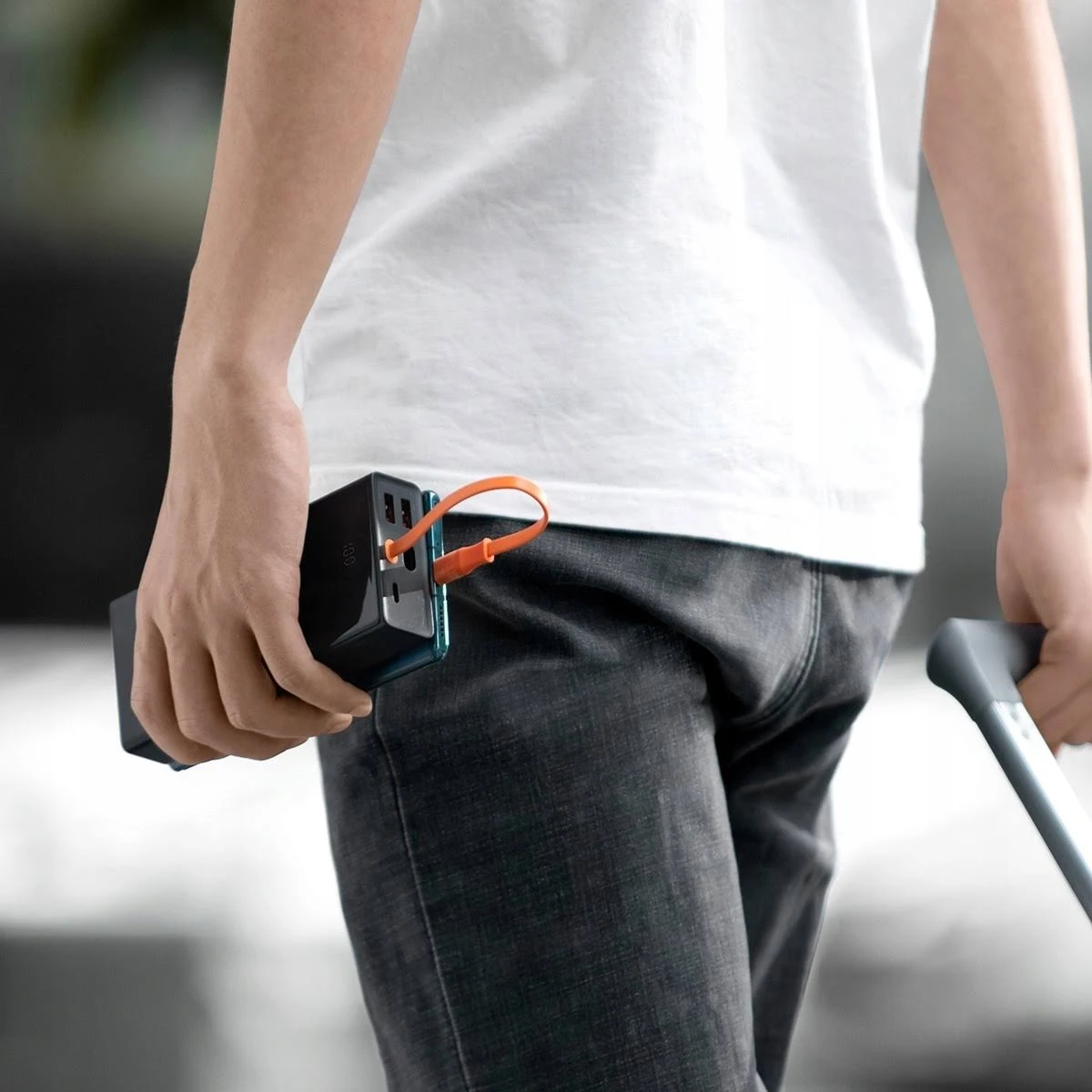 Hombre sosteniendo un cargador portátil negro con cable naranja, camisa blanca, pantalón oscuro.