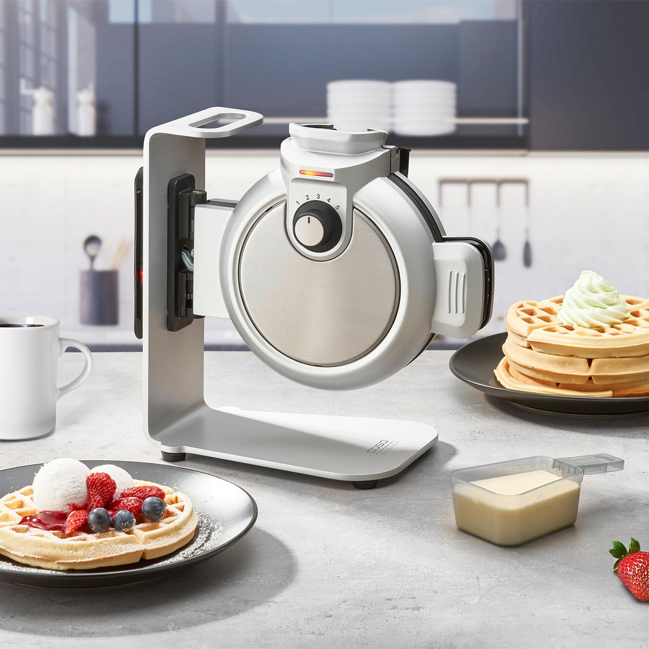 Waffeleisen mit Waffeln, Eis und Beeren auf einer Theke. Weiße Tasse links.