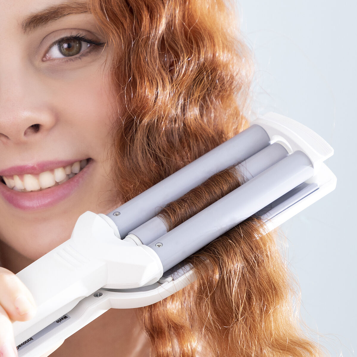 Una donna usa un arricciacapelli, con i suoi capelli rossi nello strumento, sorridendo alla fotocamera.