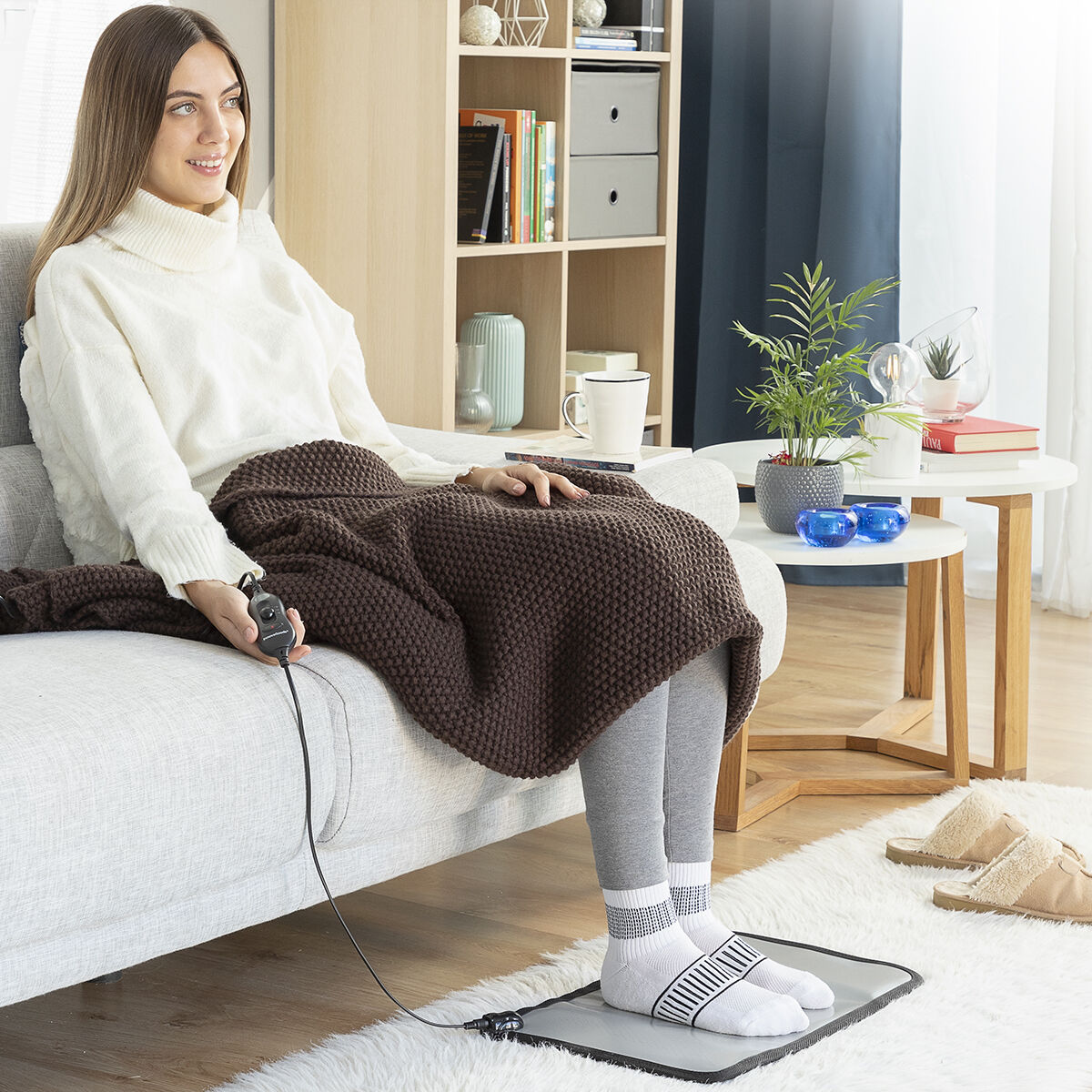 Mujer sentada en un sofá usando un calentador de pies, con una manta y decoración detrás.