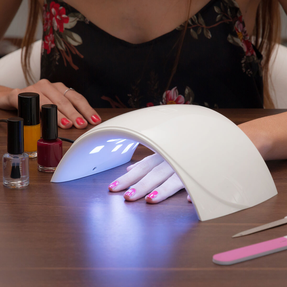 La mano de una persona bajo una lámpara de uñas blanca, con botellas de esmalte de uñas cerca sobre una mesa de madera.