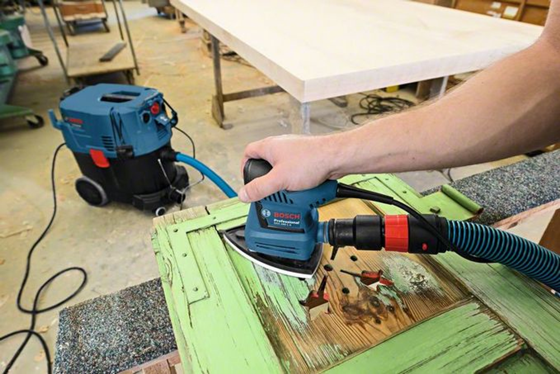 Osoba szlifująca zielone drzwi niebieską i czarną szlifierką z odkurzaczem.