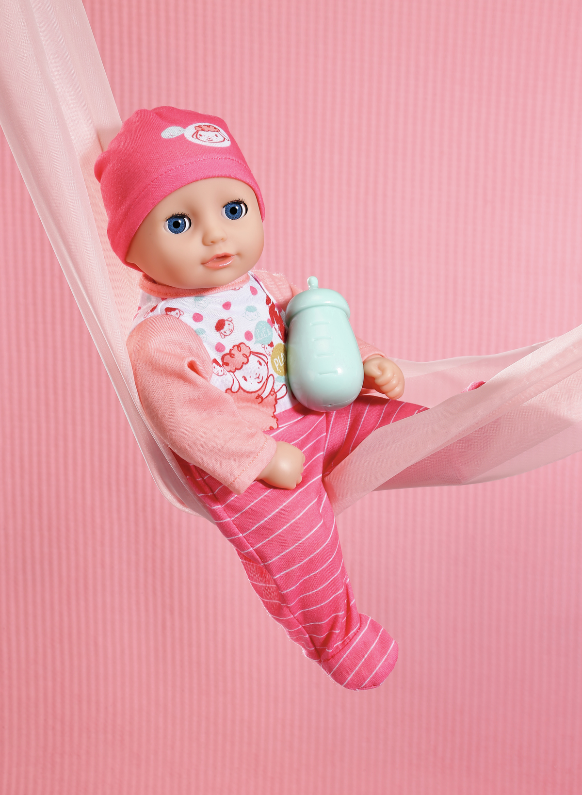 Eine Babypuppe in rosa Kleidung und Hut, die eine Flasche hält, in einer rosa Hängematte vor rosa Hintergrund liegt.