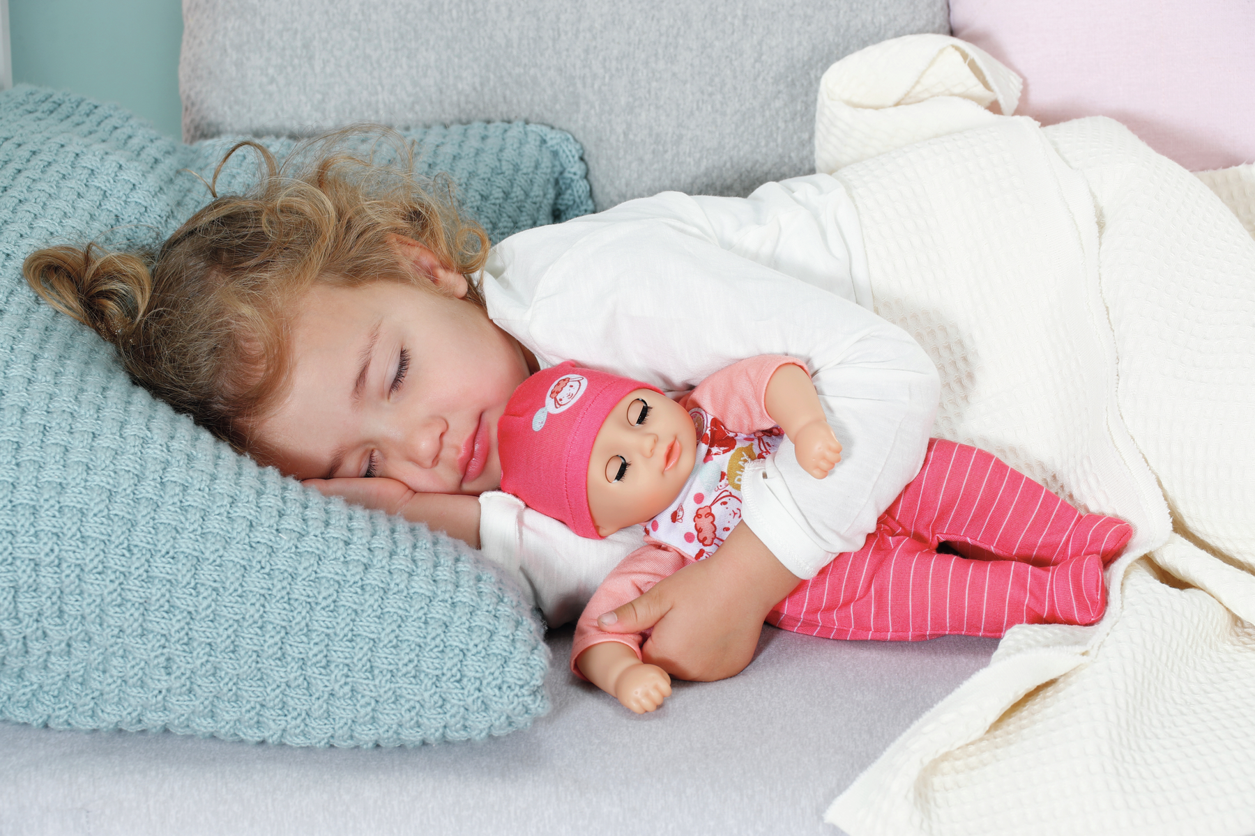 Ein junges Mädchen schläft mit einer Puppe auf einem Bett. Sie hält die Puppe eng an sich gedrückt.