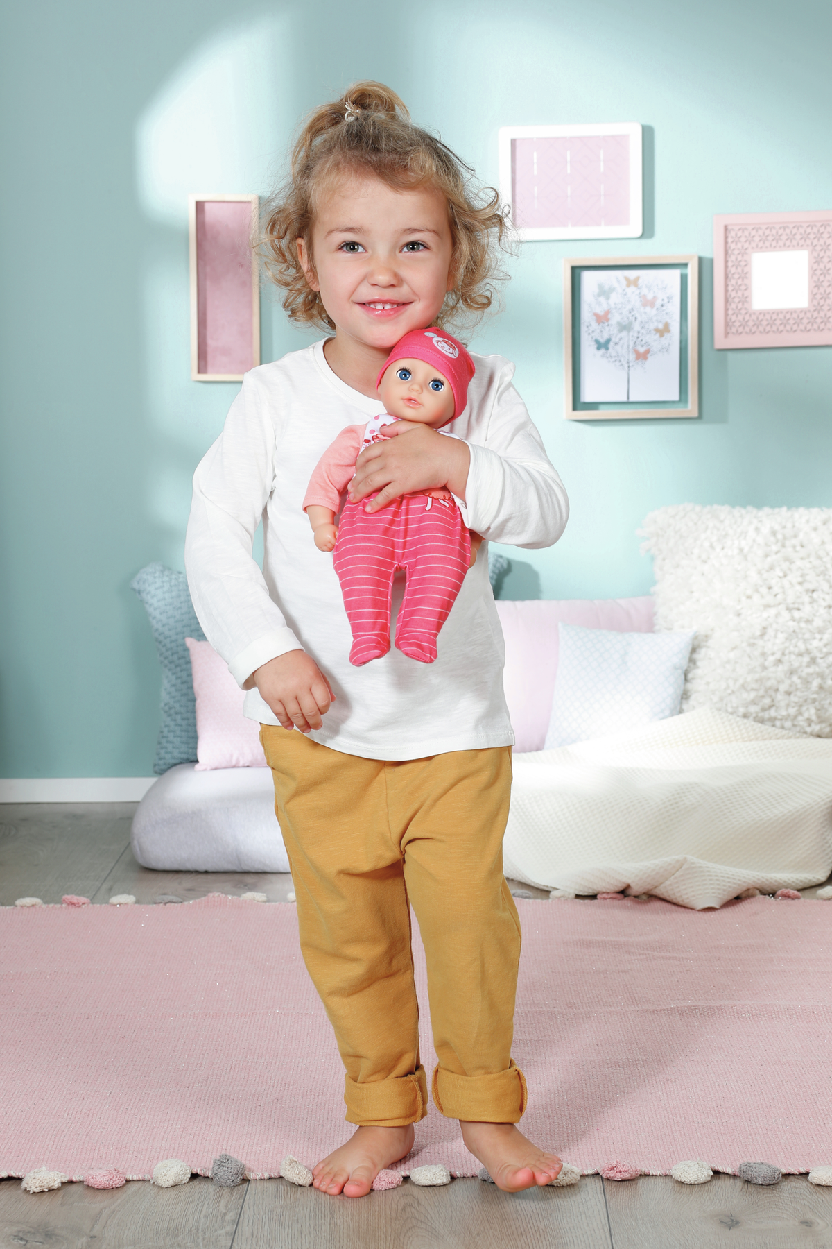 Ein junges Mädchen hält eine Puppe. Sie trägt ein weißes Hemd und eine gelbe Hose. Auf dem Boden liegt ein rosa Teppich.
