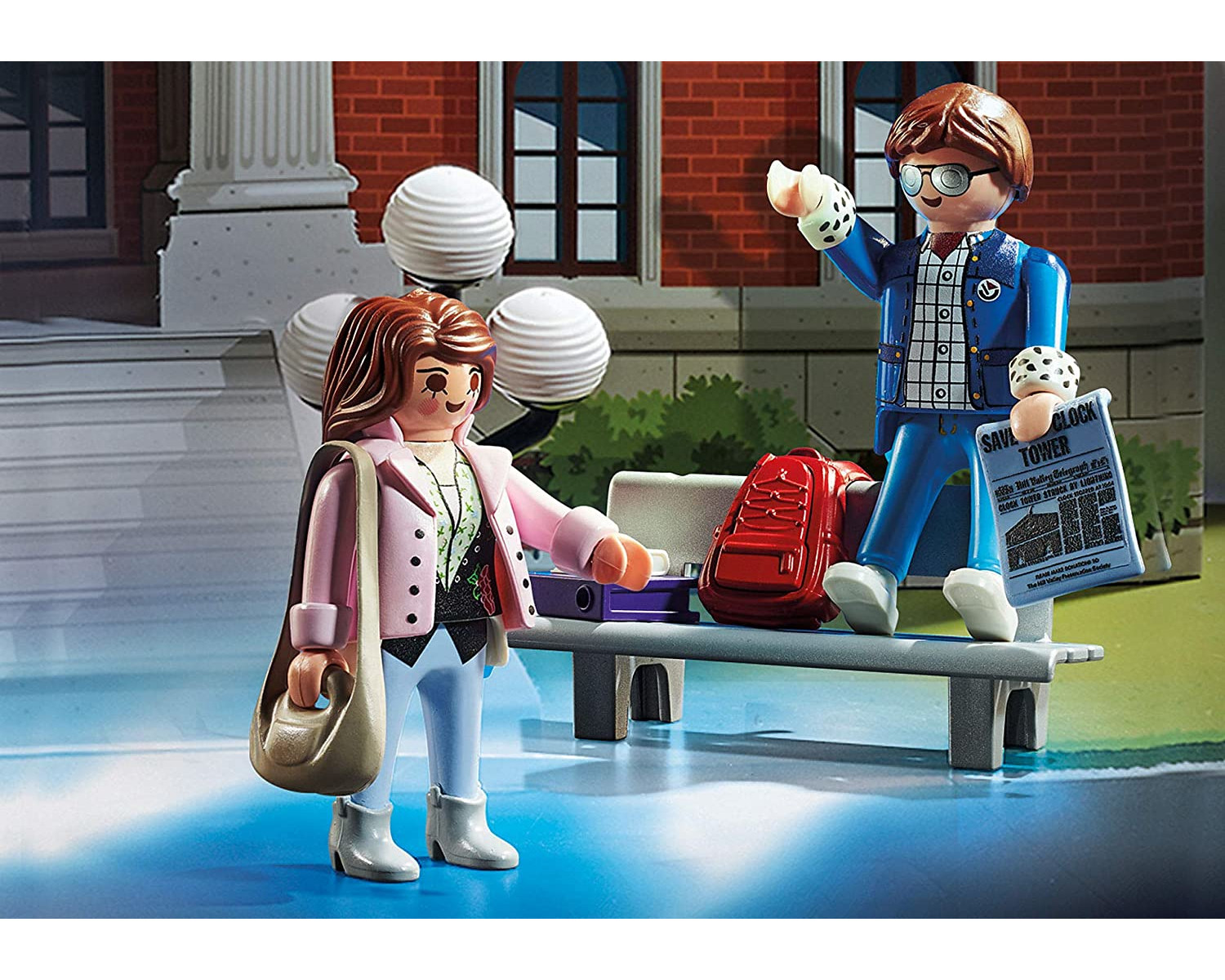 Zwei Playmobil-Figuren: eine Frau und ein Mann auf einer Bank. Szene hat einen roten Rucksack und ein Gebäude.