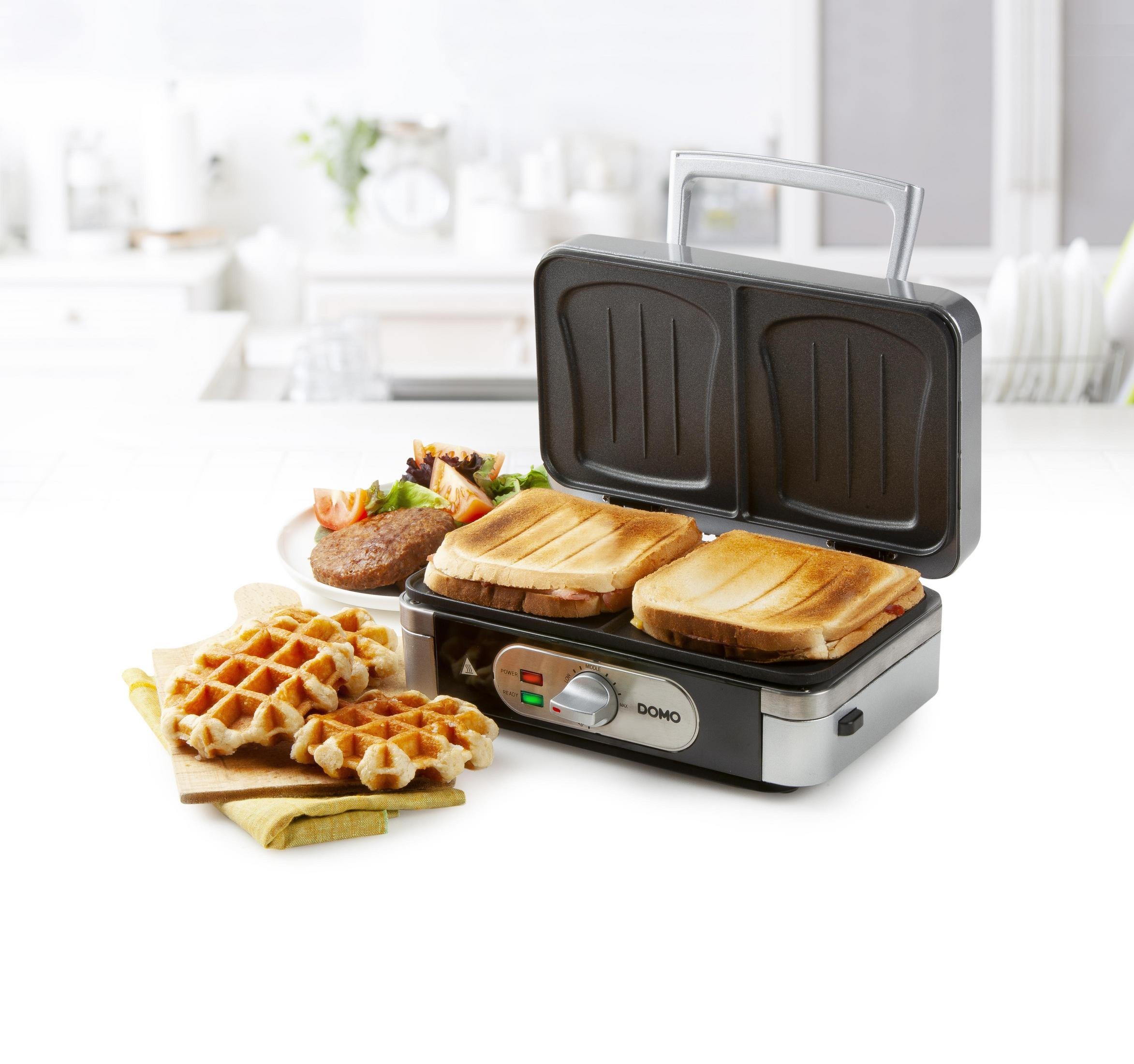 Toaster mit zwei Sandwiches, zwei Waffeln und anderen Speisen. Es steht auf einem weißen Tisch in einer Küche.