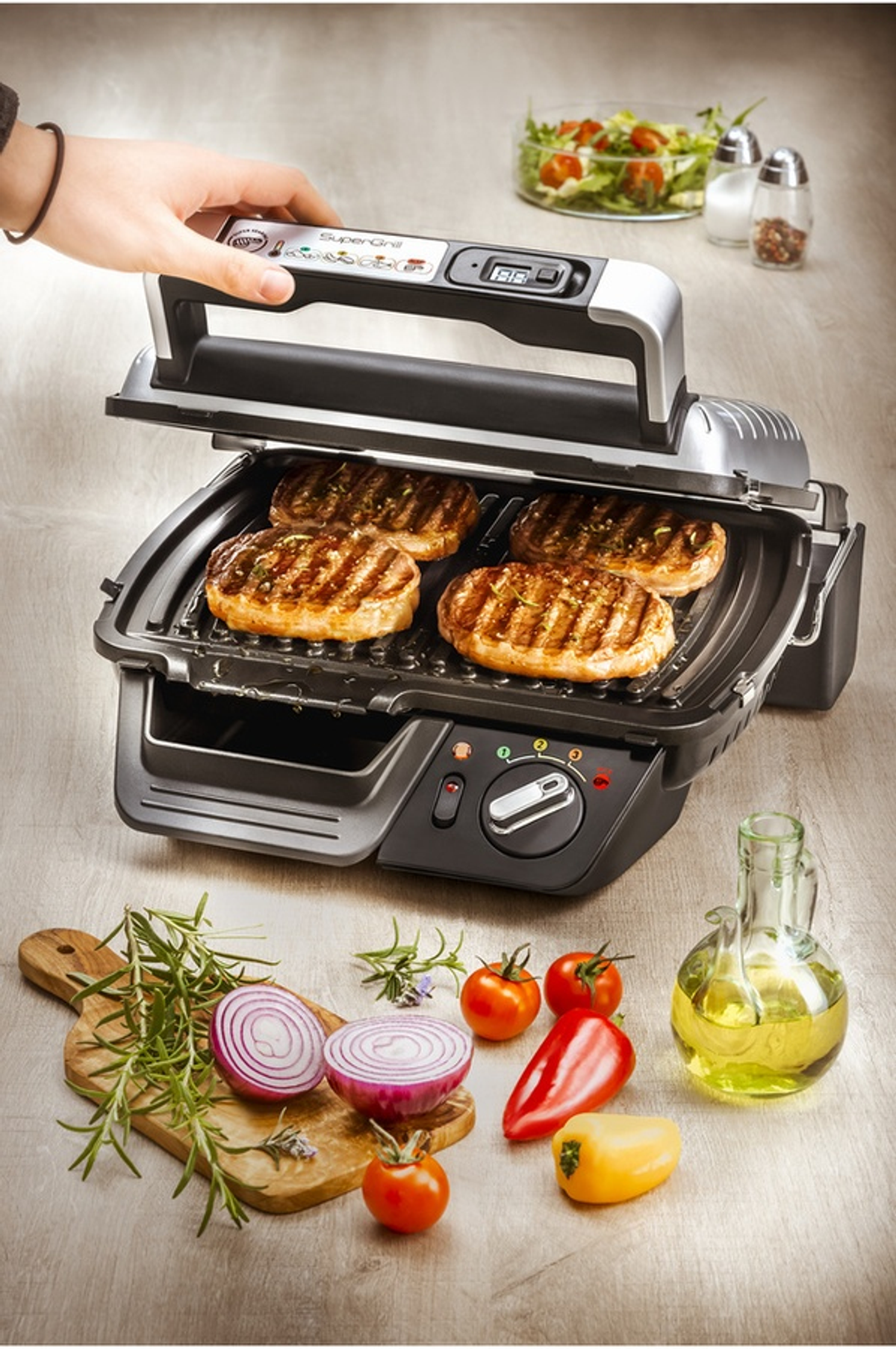 Parrilla con cuatro filetes. La parrilla está abierta. Tiene tomates rojos y otros alimentos alrededor.