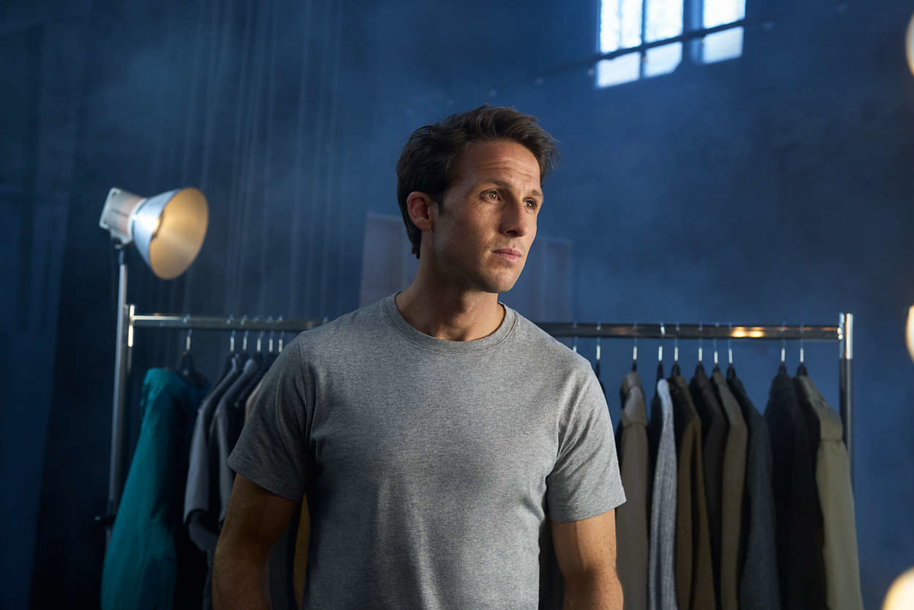 Mężczyzna w szarej koszulce stoi przed wieszakiem na ubrania i światłami studyjnymi.