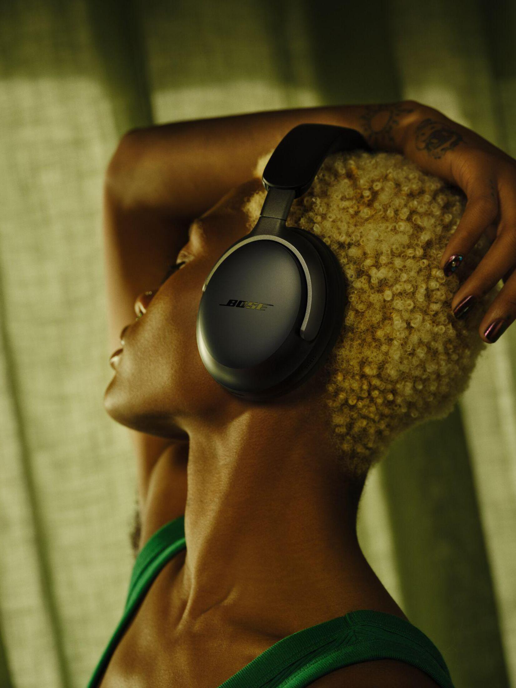 Kobieta z krótkimi, kręconymi blond włosami nosząca czarne słuchawki Bose i zielony top.
