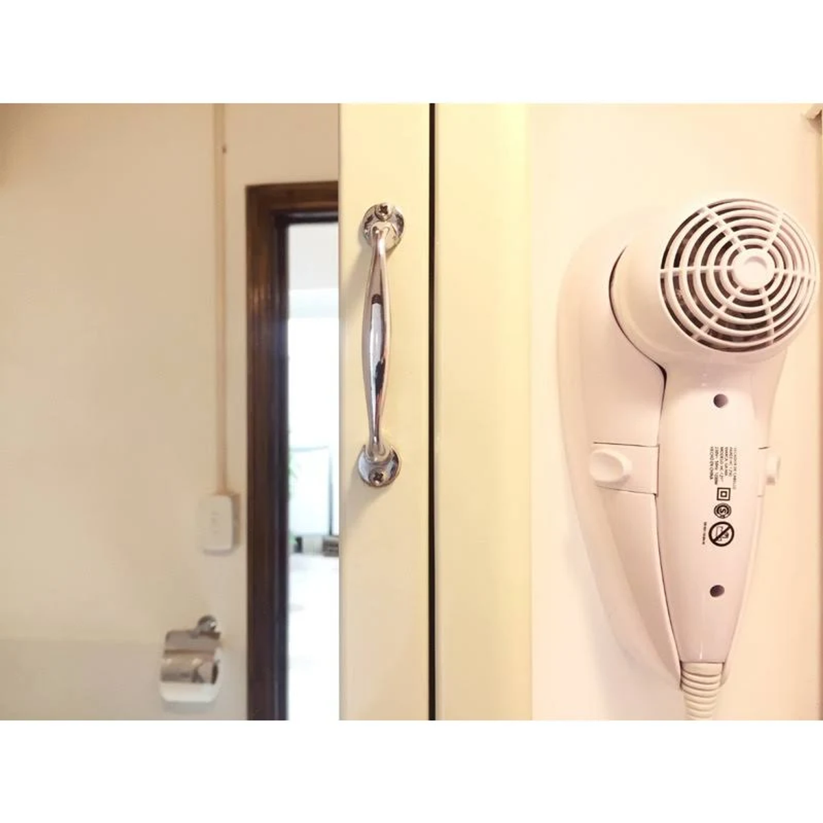 Biała suszarka do włosów zamontowana na ścianie, klamka na drzwiach, lustro i uchwyt na papier toaletowy.