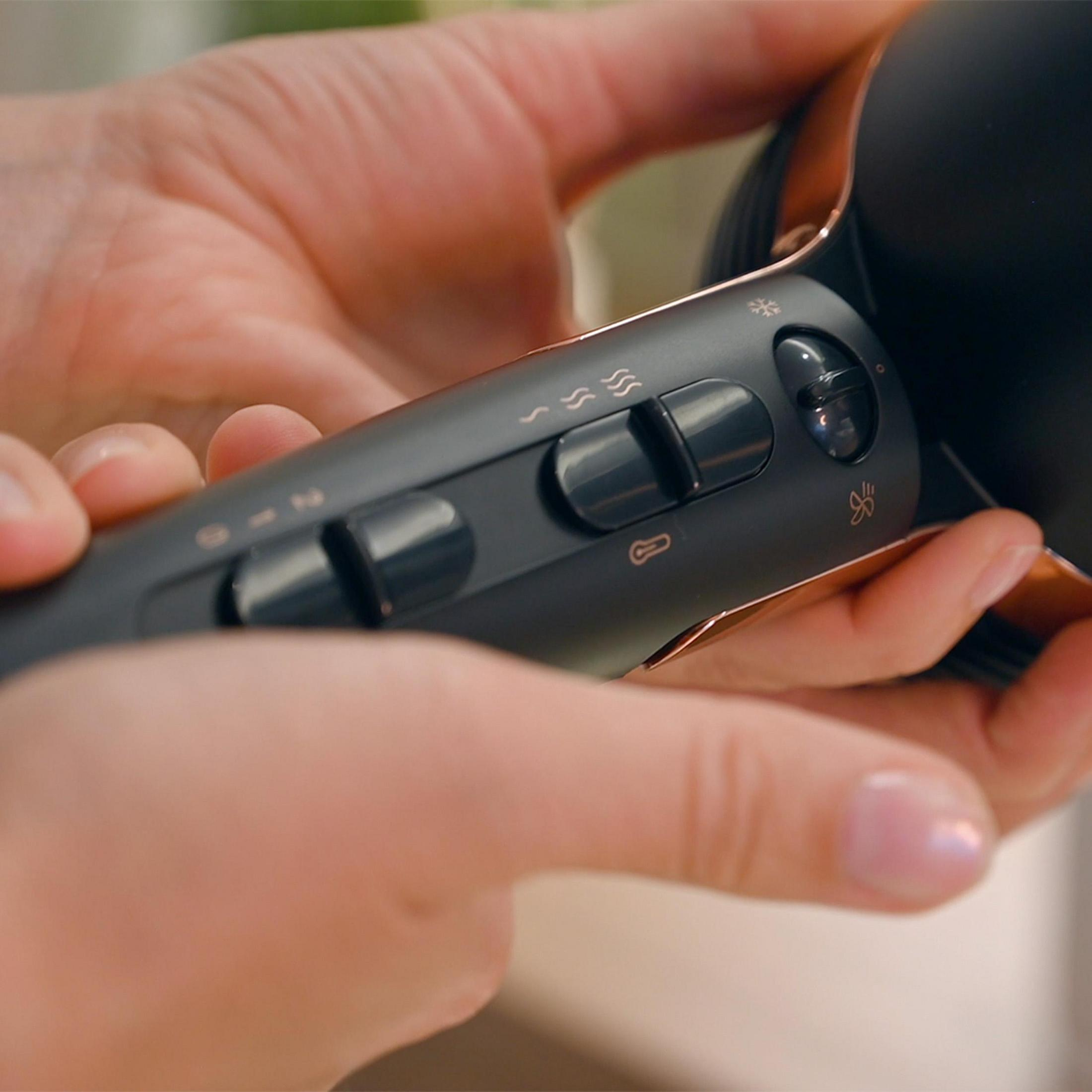 Dłonie trzymające czarną i miedzianą suszarkę do włosów, pokazujące ustawienia sterowania.